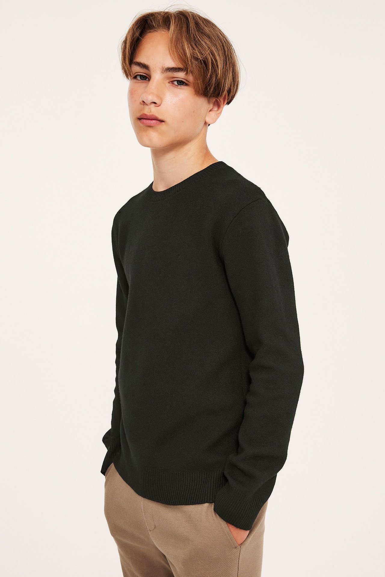 Widok z przodu dziecka w czarnym, dzianinowym swetrze z okrągłym dekoltem, ściągaczami i beżowych spodniach.