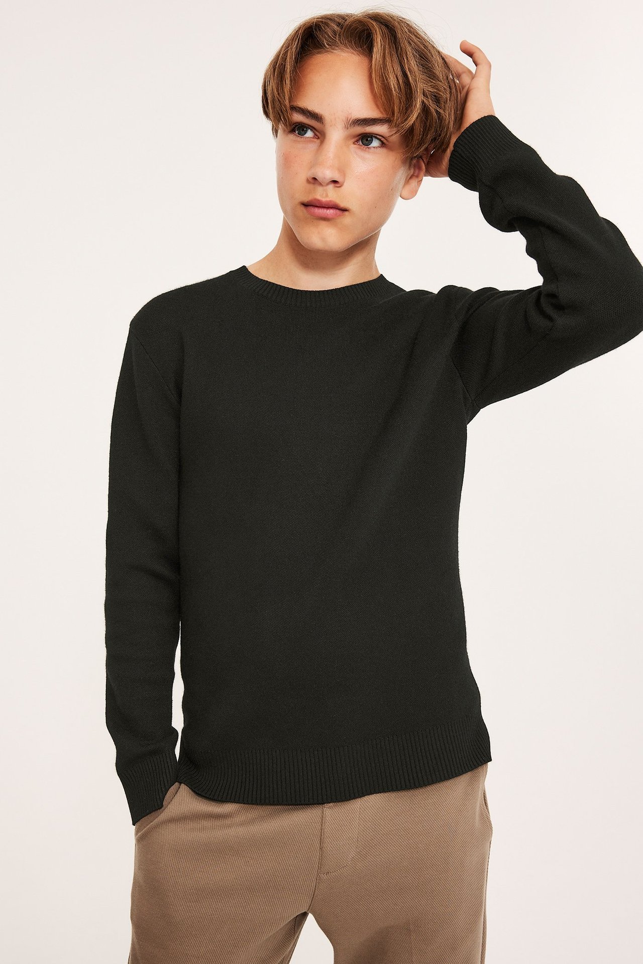 Widok z przodu: dziecko w czarnym, dzianinowym swetrze z okrągłym dekoltem i beżowych spodniach.