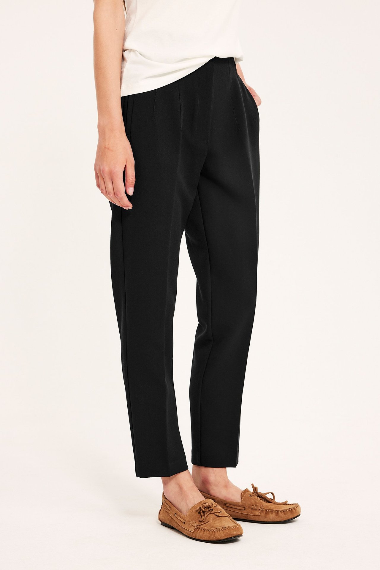 Sidebilde av kvinne i svarte smale høy midje bukser, hvit t-skjorte og brune mokkasiner.