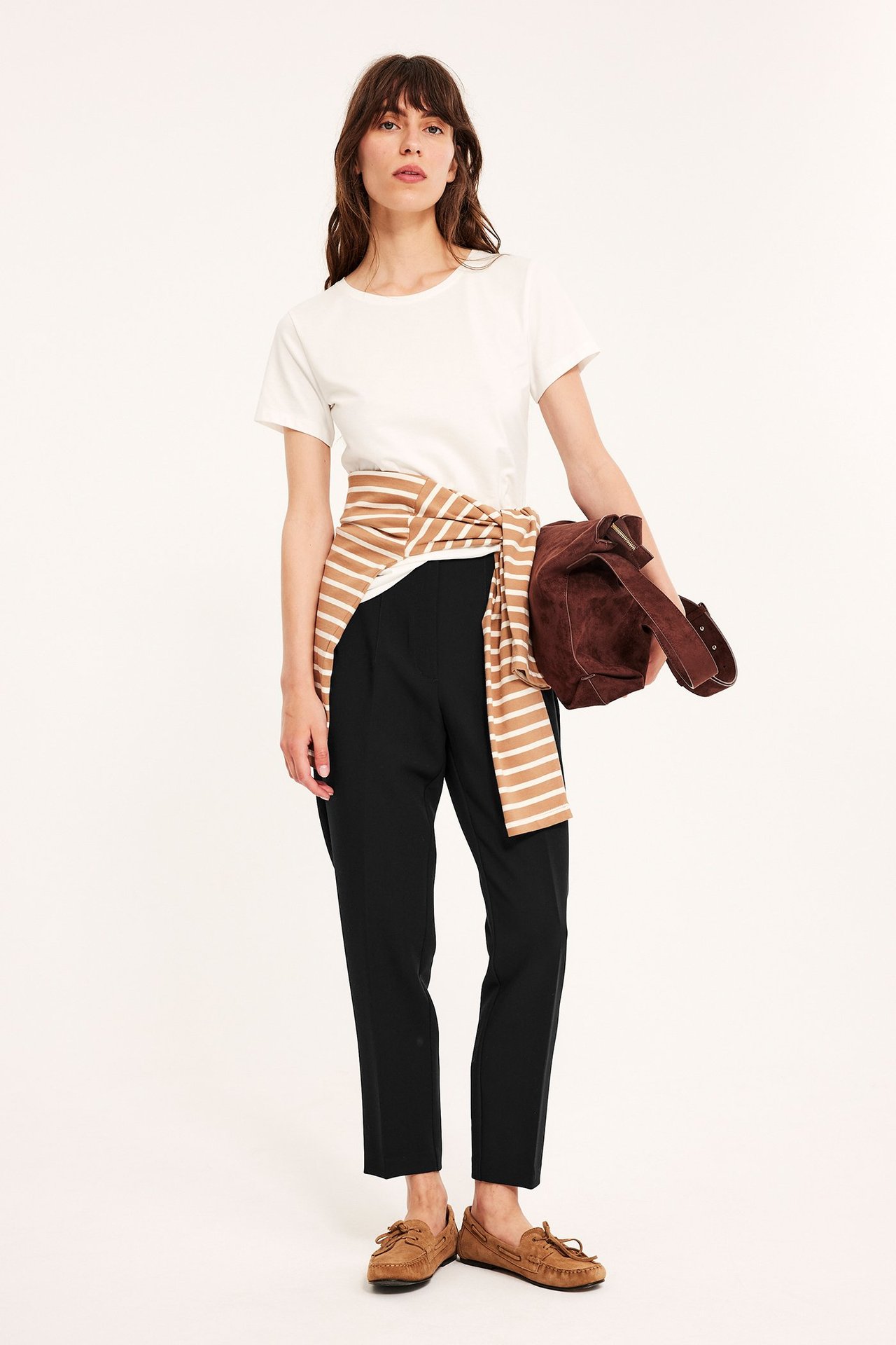 Widok z przodu: Kobieta w czarnych, wąskich spodniach, białym t-shircie, pasiastym topie, brązowych mokasynach, zamszowej torbie.