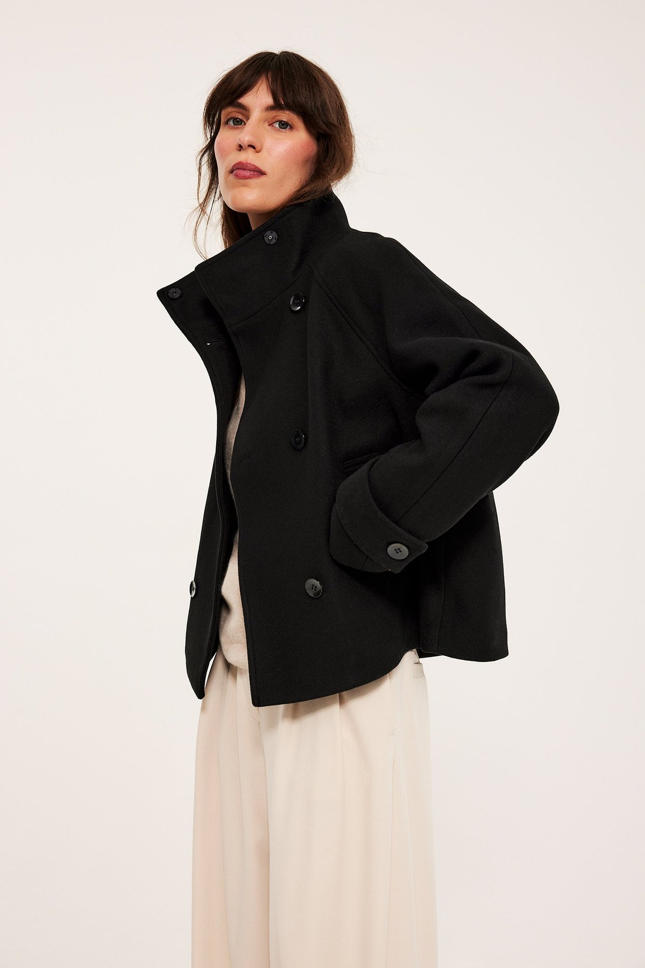 Edestä: nainen mustassa lyhyessä kaksirivisessä takissa korkealla kauluksella, vaaleanbeigessä topissa ja leveälahkeisissa housuissa.