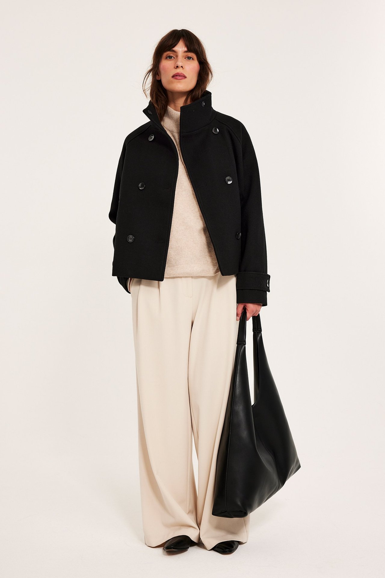 Nainen yllään musta kaksirivinen takki, beige neule, vaaleanbeiget housut ja pitää mustaa kangaskassia.