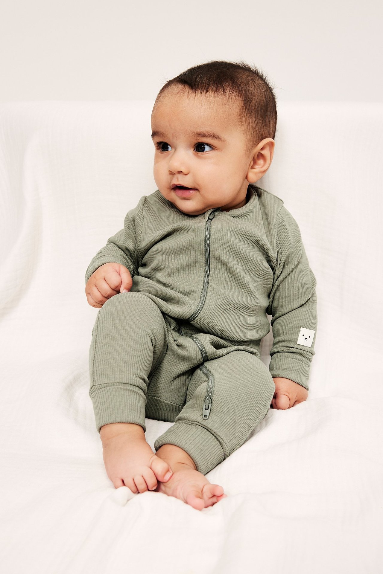 Framifrån: Baby i mörkgrön ribbad jumpsuit med tvåvägsdragkedja, uppvikta muddar och björnmärke.