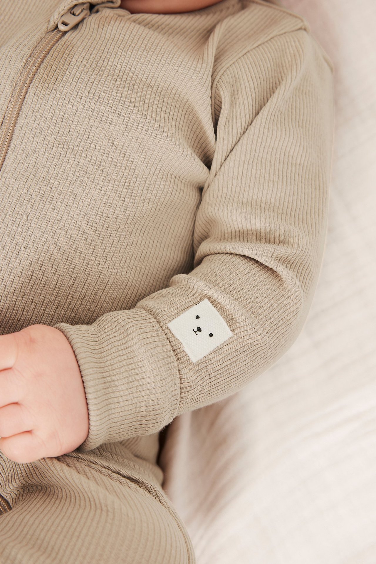 Närbild på baby i beige ribbad bomullsjumpsuit med tvåvägsdragkedja och björnapplikation på ärmen.