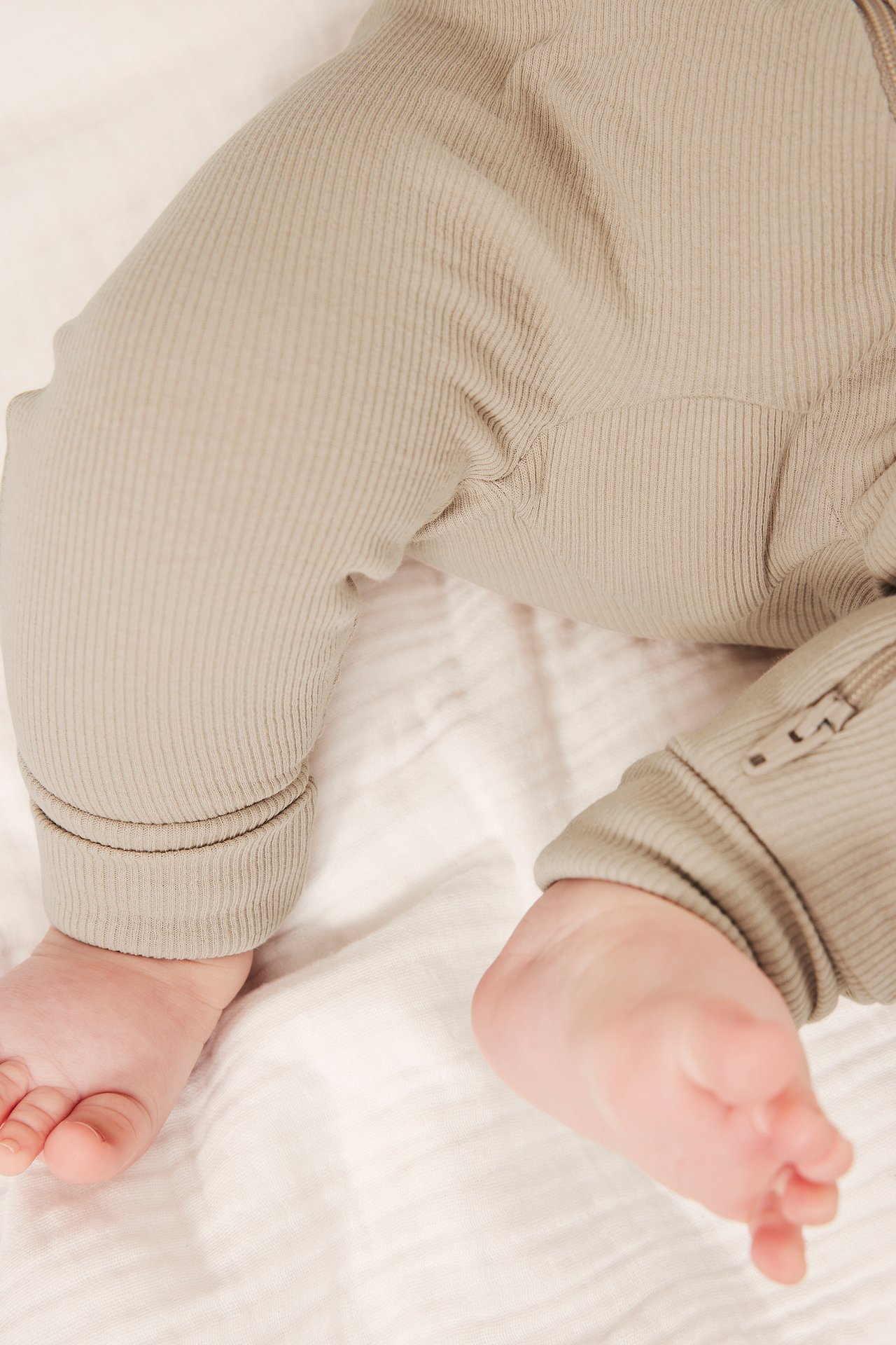 Nærbilde av baby i beige ribbet ensfarget jumpsuit med oppbrettede mansjetter og delvis glidelås.