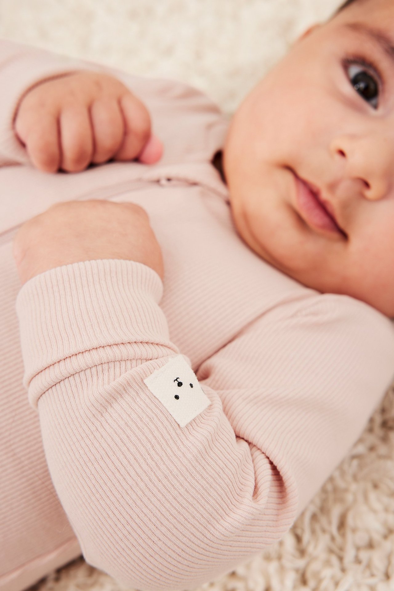Närbild på en baby i en rosa ribbad jumpsuit med vikta muddar och liten björnetikett.
