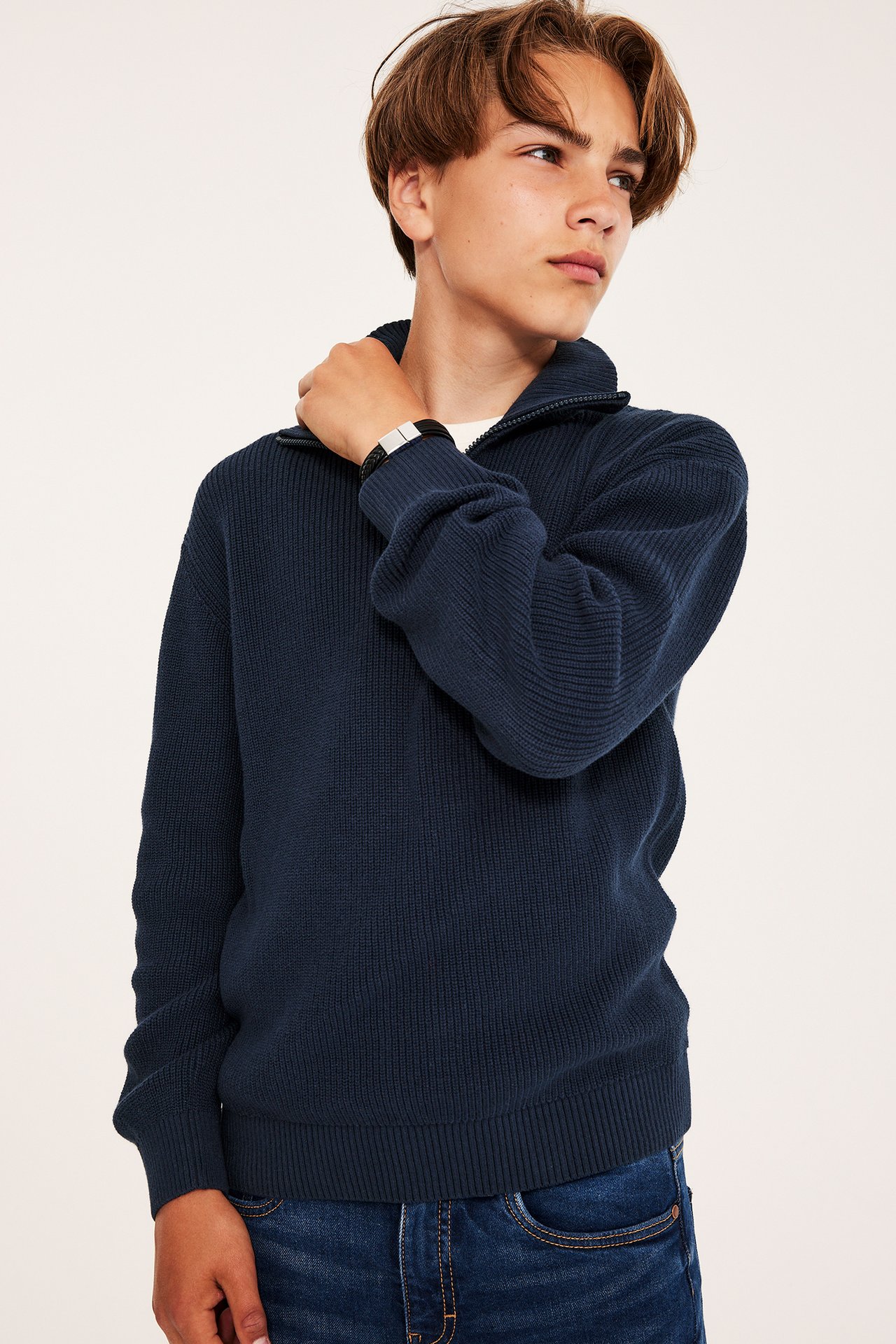 Frontvy av en kid i mörkblå stickad bomullströja med halv dragkedja, blå jeans och svart armband.