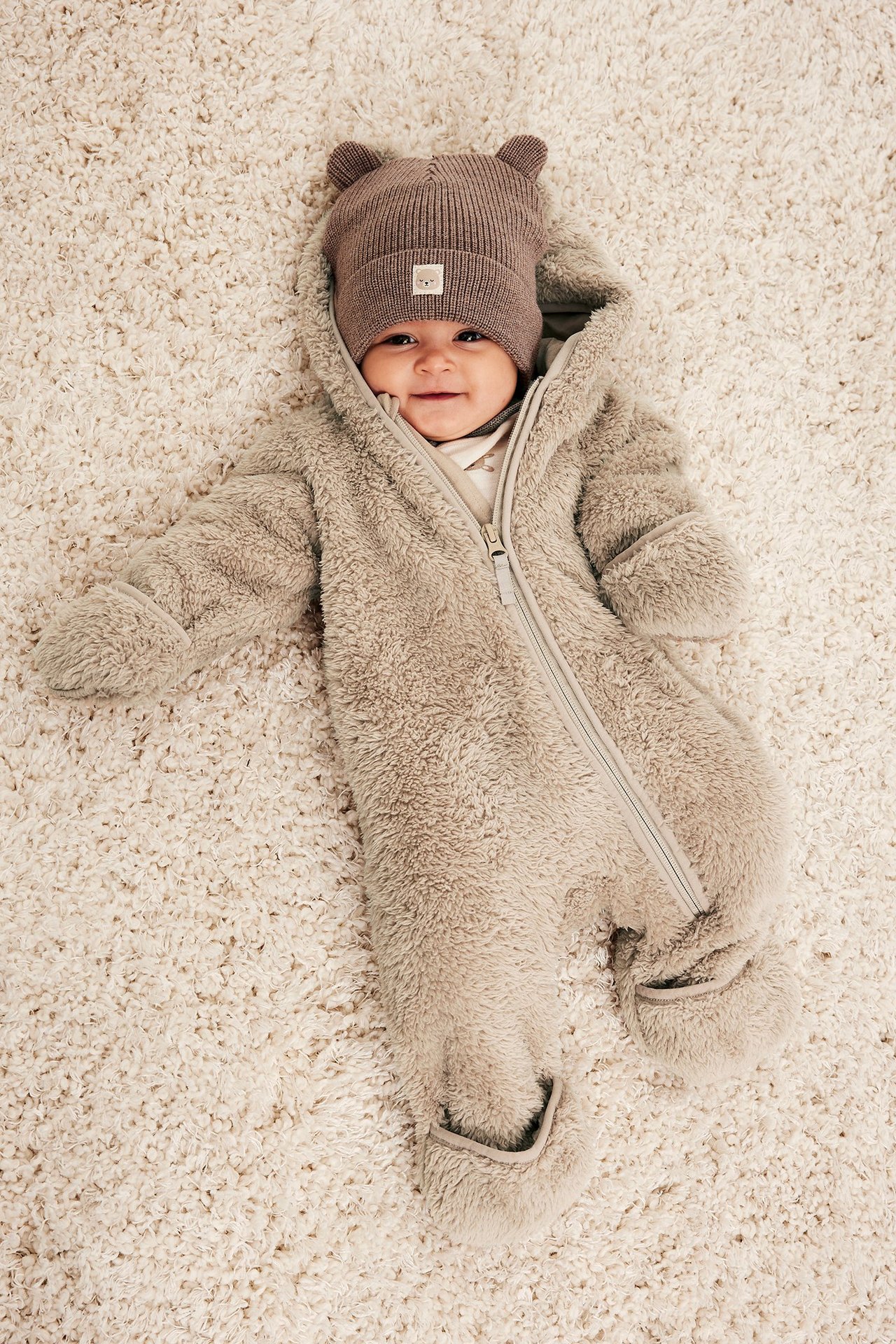 Ovanifrån, en baby i beige pileoverall med dragkedja och en brun ribbad mössa med öron.