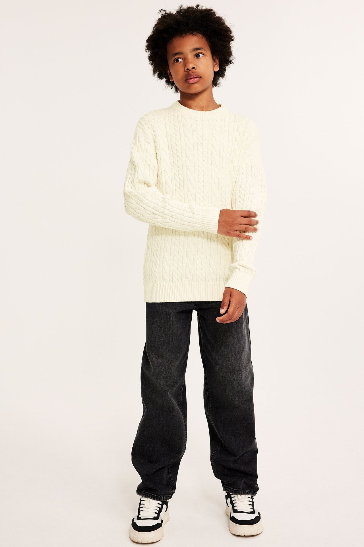 Widok z przodu dziecka w kremowym swetrze z warkoczowym splotem, czarnych szerokich dżinsach i białych trampkach.