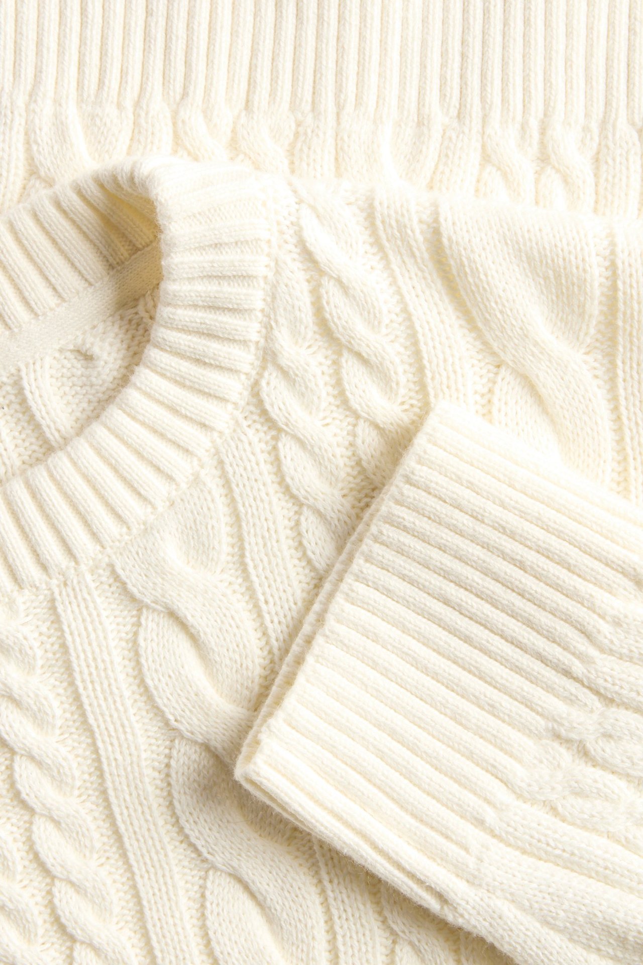 Zbliżenie na kremowy bawełniany sweter w warkocze z prążkowanym dekoltem i mankietem.