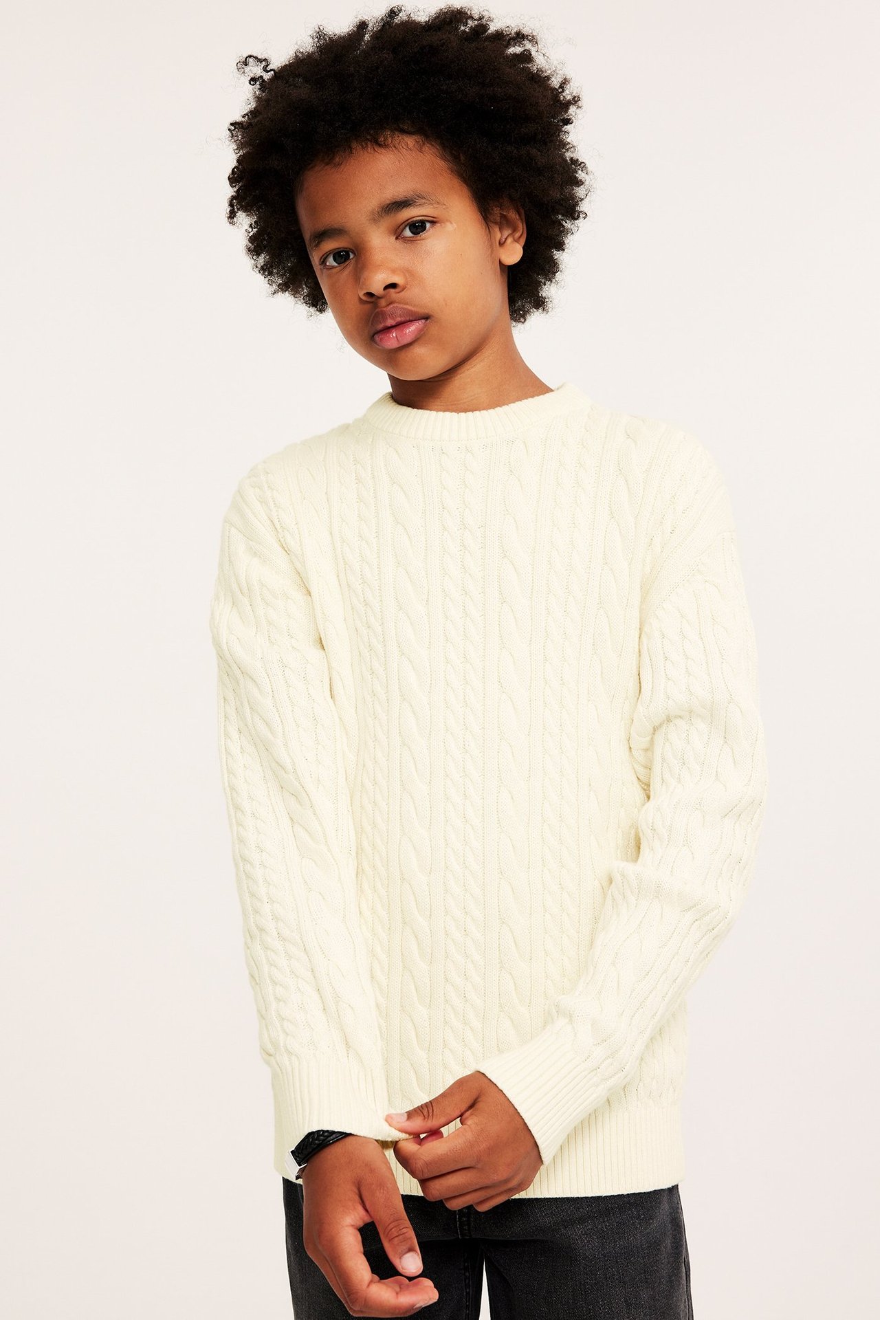 Widok z przodu dziecka w kremowym bawełnianym swetrze w warkocze i ciemnych dżinsach.