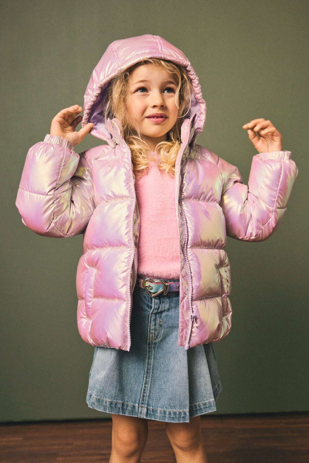 Framifrån: Barn i skimrande rosa pufferjacka, rosa tröja och jeanskjol.