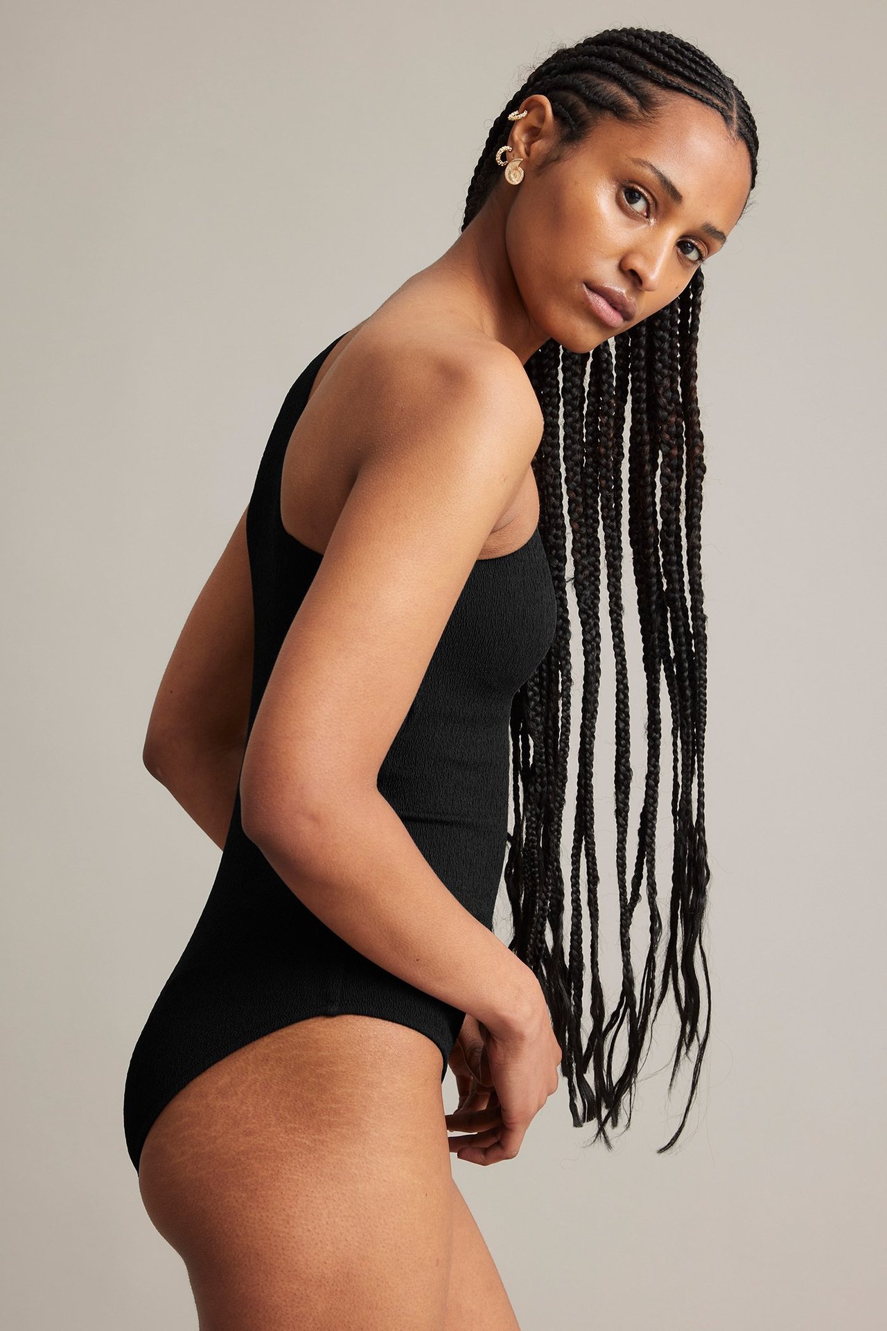 Sidebilde av en kvinne i svart enskuldret badedrakt med struktur, gulløreringer og ørepynt.