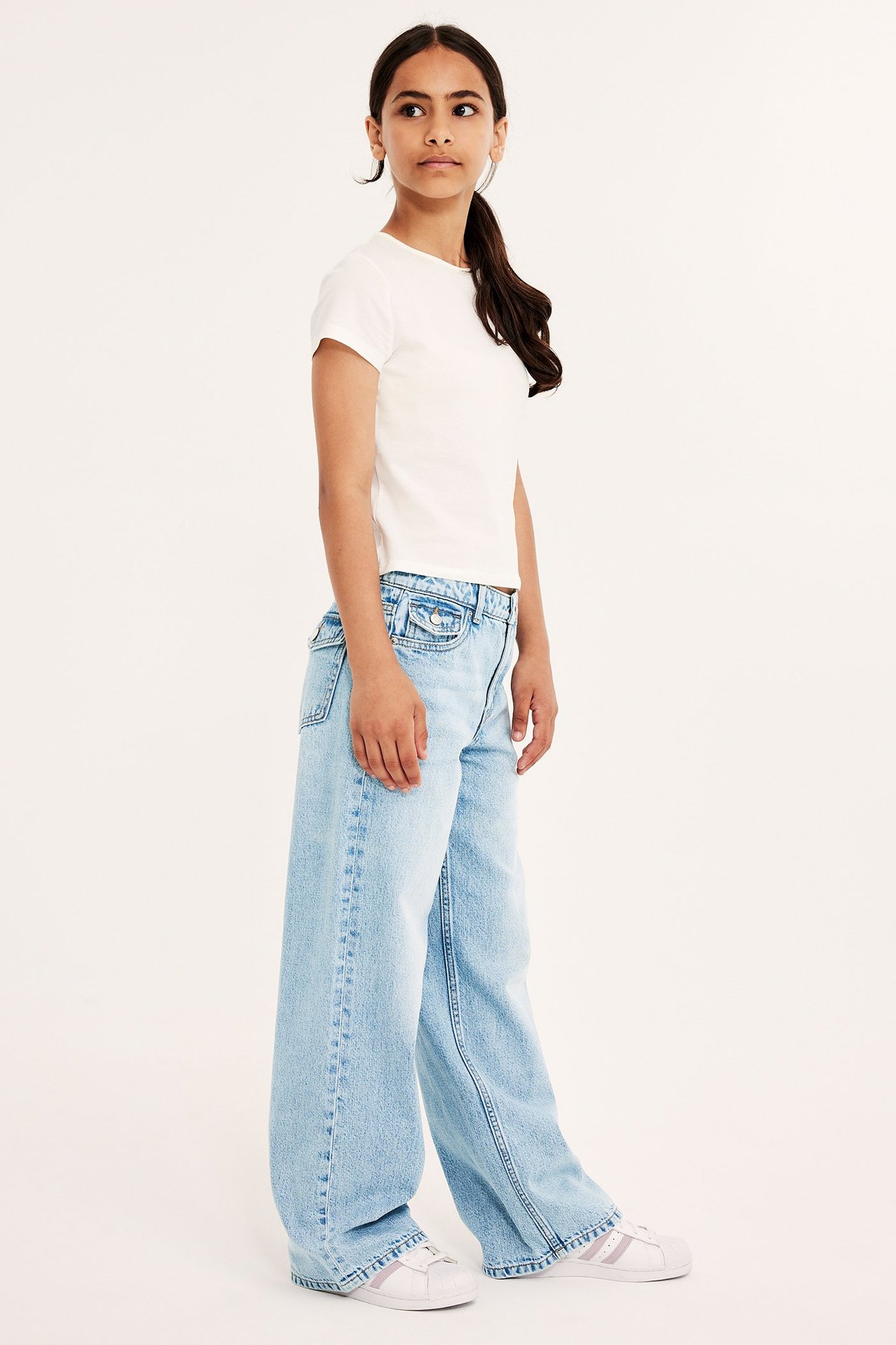 Dziewczynka w białej koszulce i luźnych jasnoniebieskich jeansach z niskim stanem, ozdobne kieszenie.