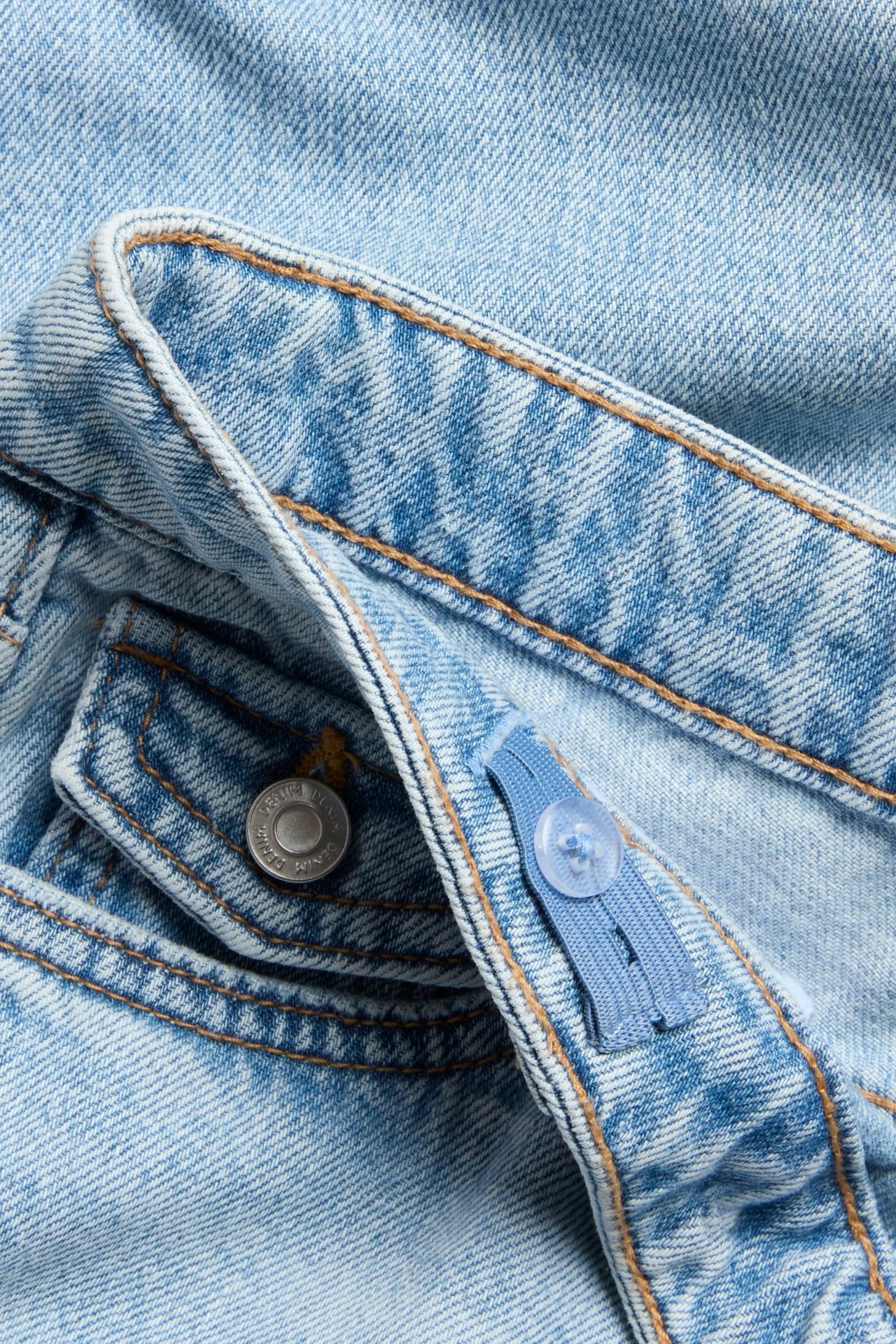 Zbliżenie na jasnoniebieskie luźne jeansy z denimu, brązowe szwy, srebrny guzik, regulowana gumka w pasie.