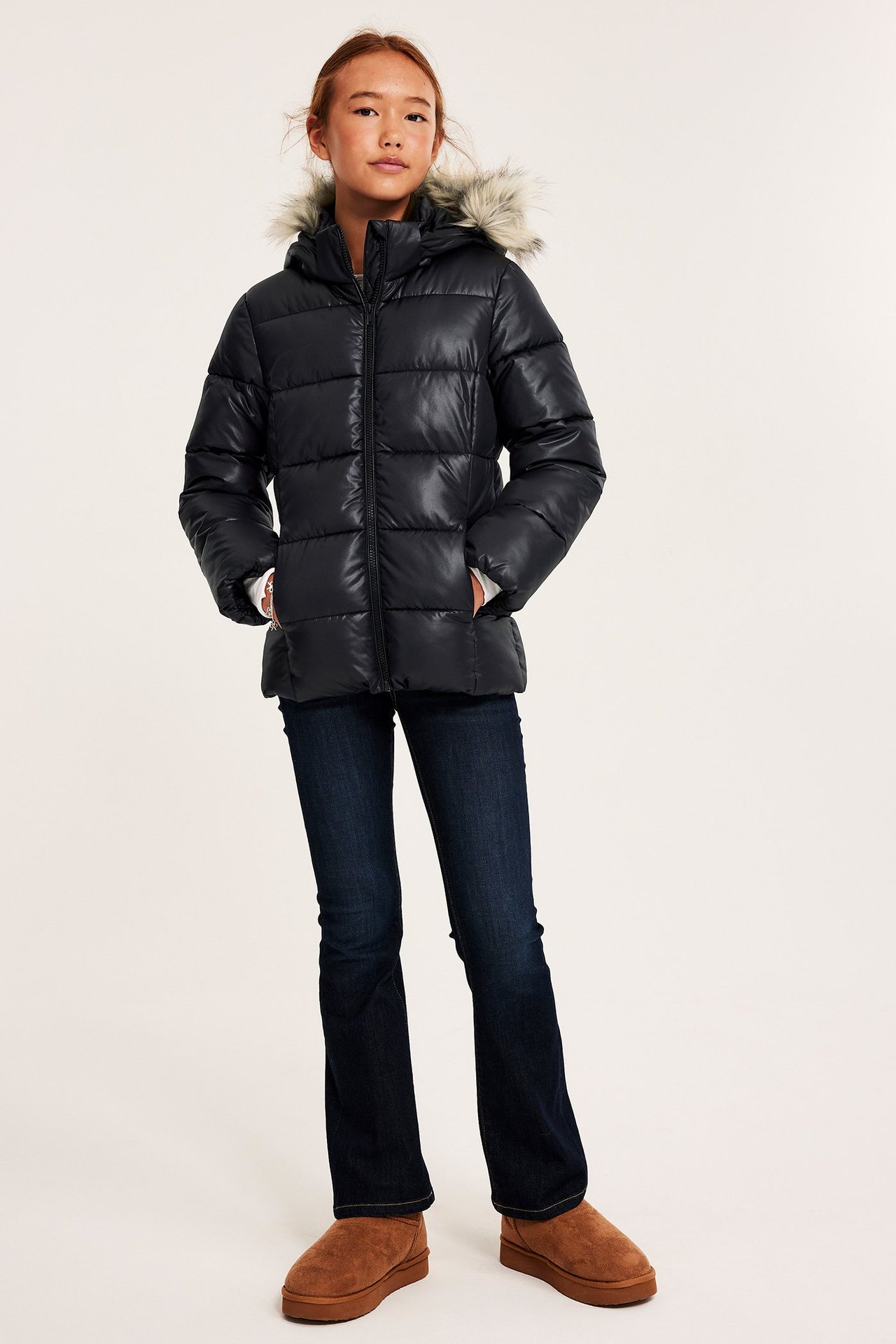 Frontvy av ett barn i svart pufferjacka med fuskpäls på huvan, mörkblå jeans och bruna ankelboots.