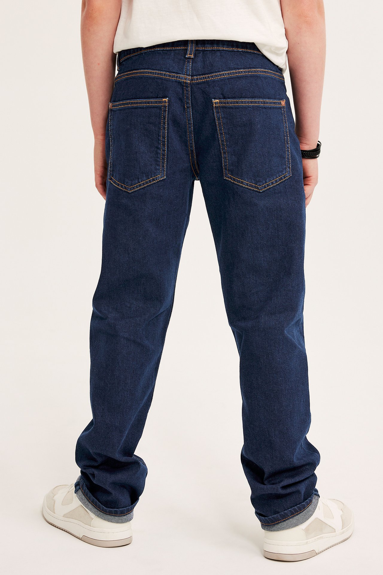 Bakfra på et barn i mørkeblå rette jeans med oransje sømmer, en hvit t-skjorte og hvite joggesko.