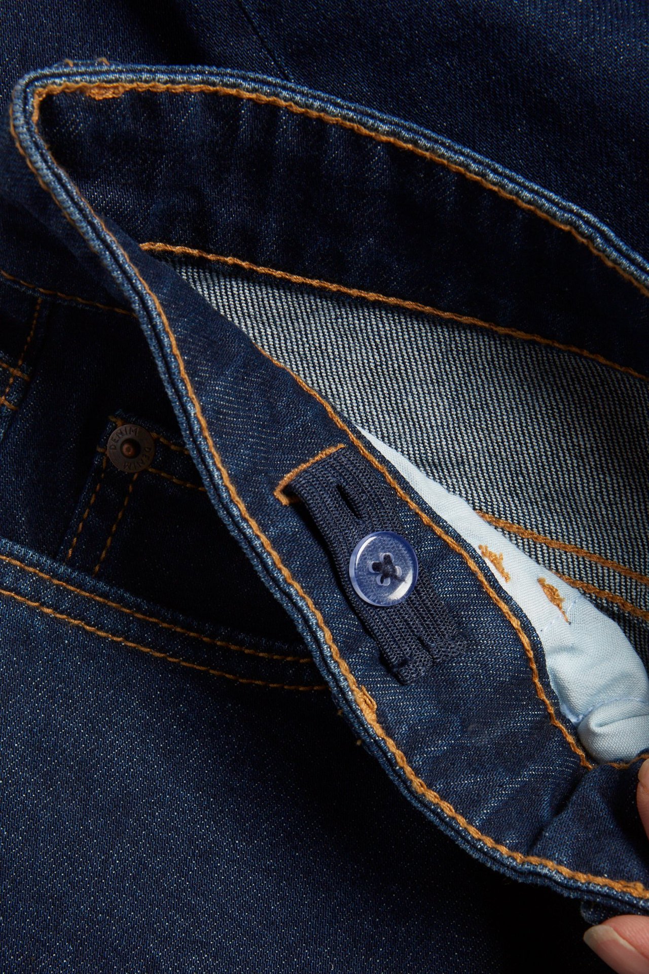 Zbliżenie na pas ciemnoniebieskich prostych dżinsów z regulowaną gumką i guzikiem, z pomarańczowymi szwami.