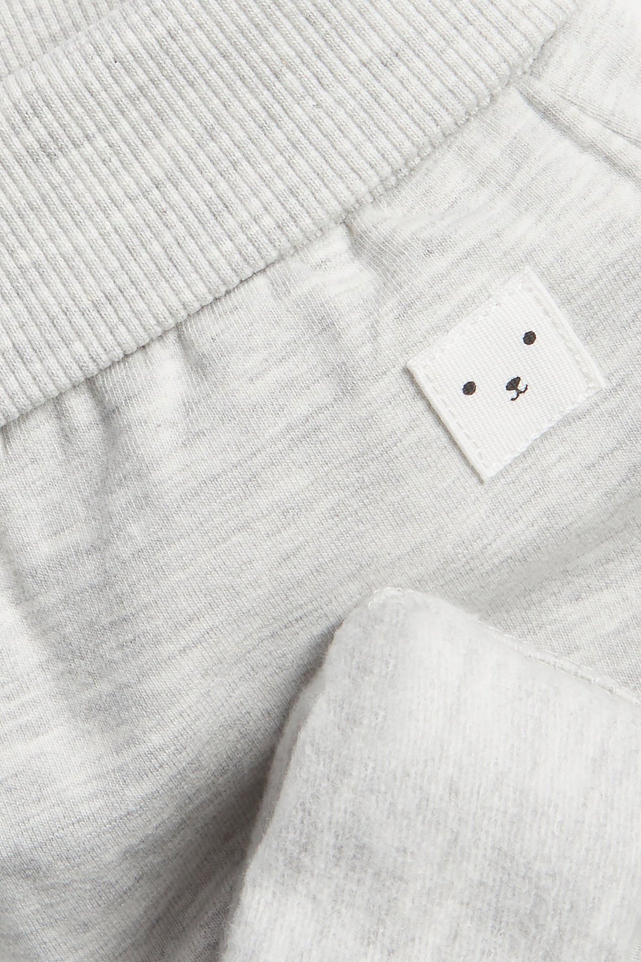 Zbliżenie jasnoszarych melanżowych spodni dresowych dla niemowląt z prążkowanym pasem i metką z misiem.