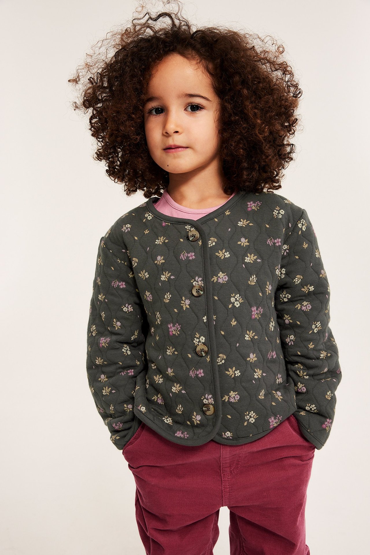 Barn i en mørkegrå blomstret quiltet jakke med knapper, over en rosa t-skjorte og røde kordfløyelsbukser.
