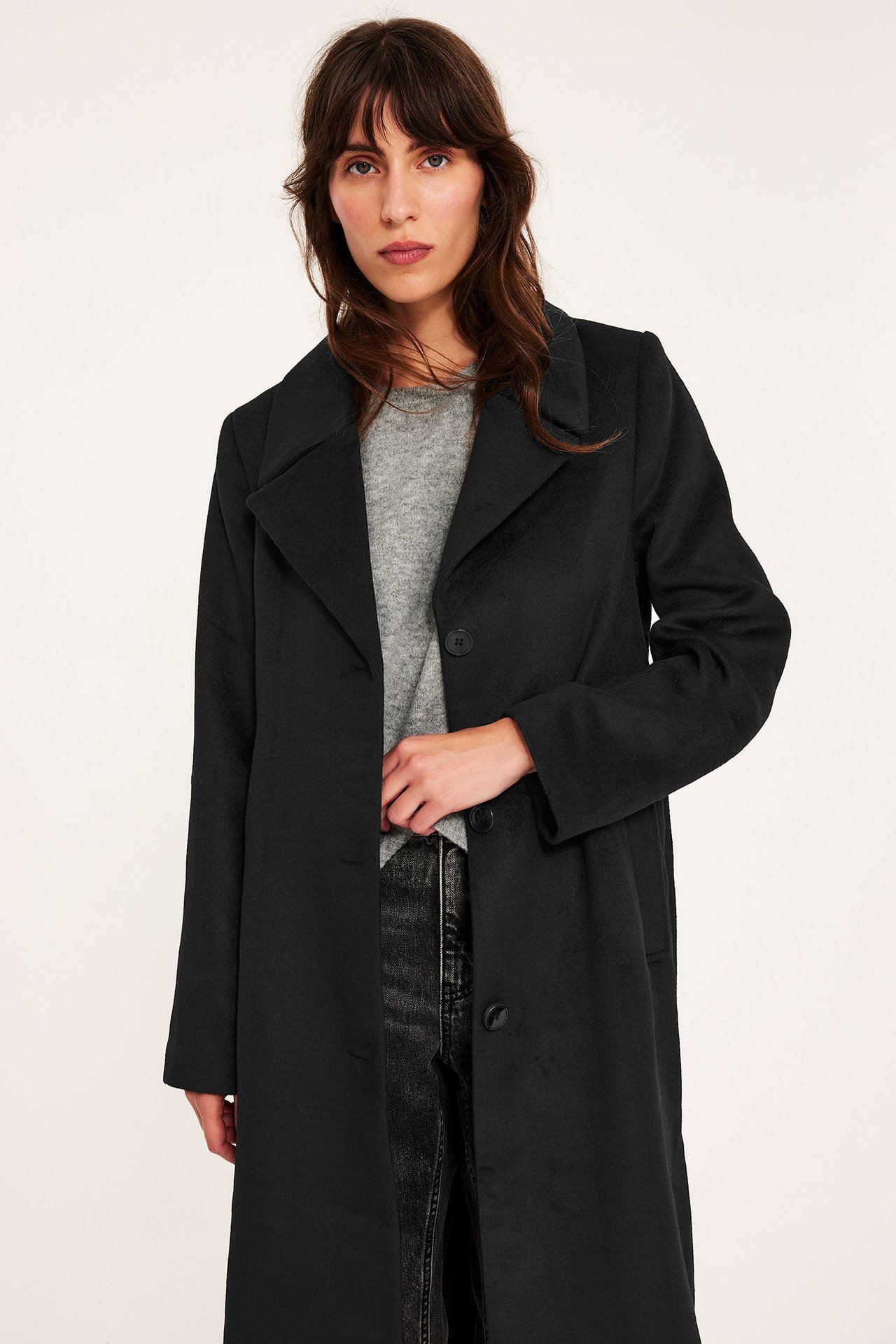 Edestä näkymä naisesta, jolla on avoin pitkä musta takki harmaan neuleen ja tummien farkkujen päällä.