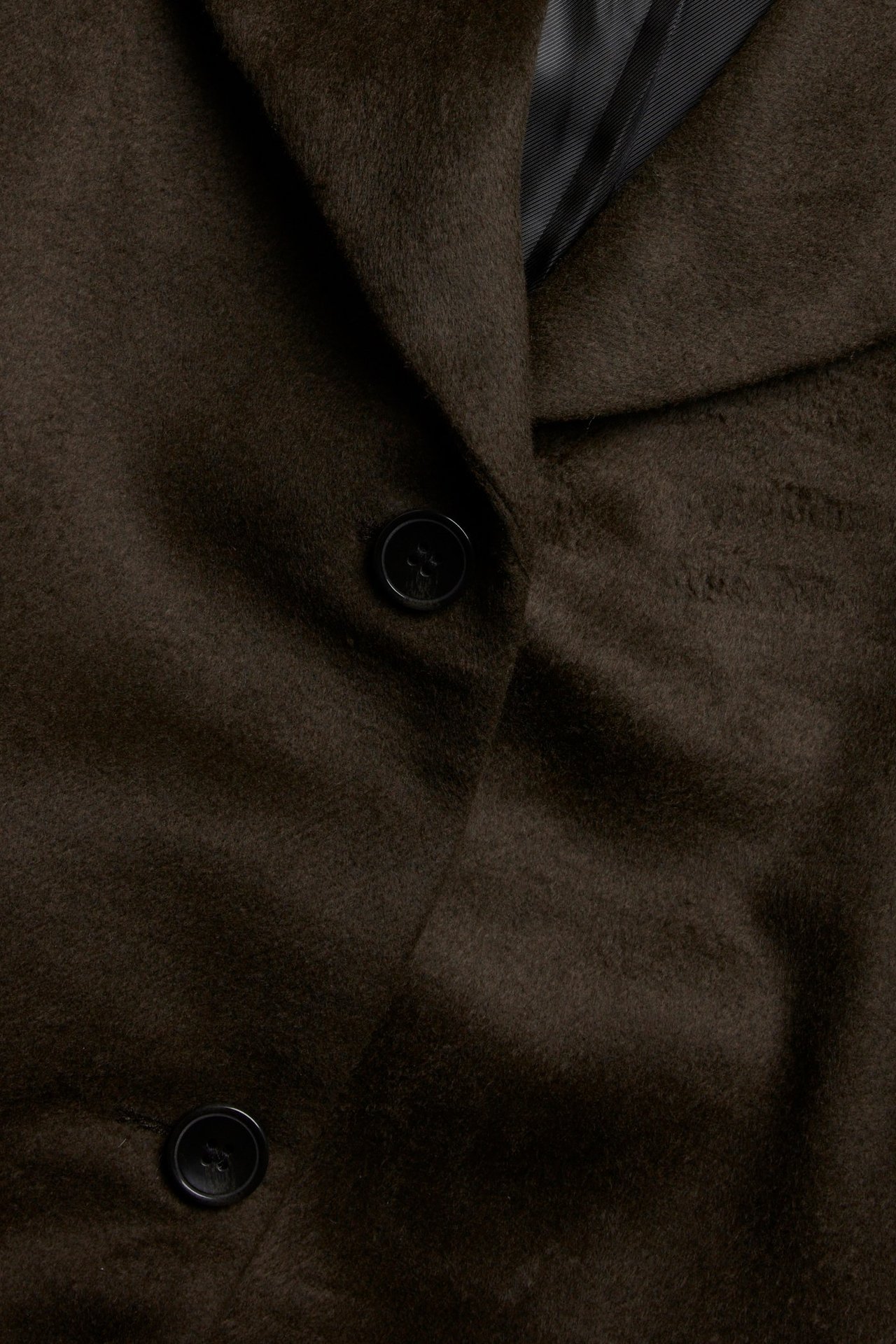 Zbliżenie brązowego wełnianego płaszcza z dwoma czarnymi guzikami i czarną podszewką w paski.