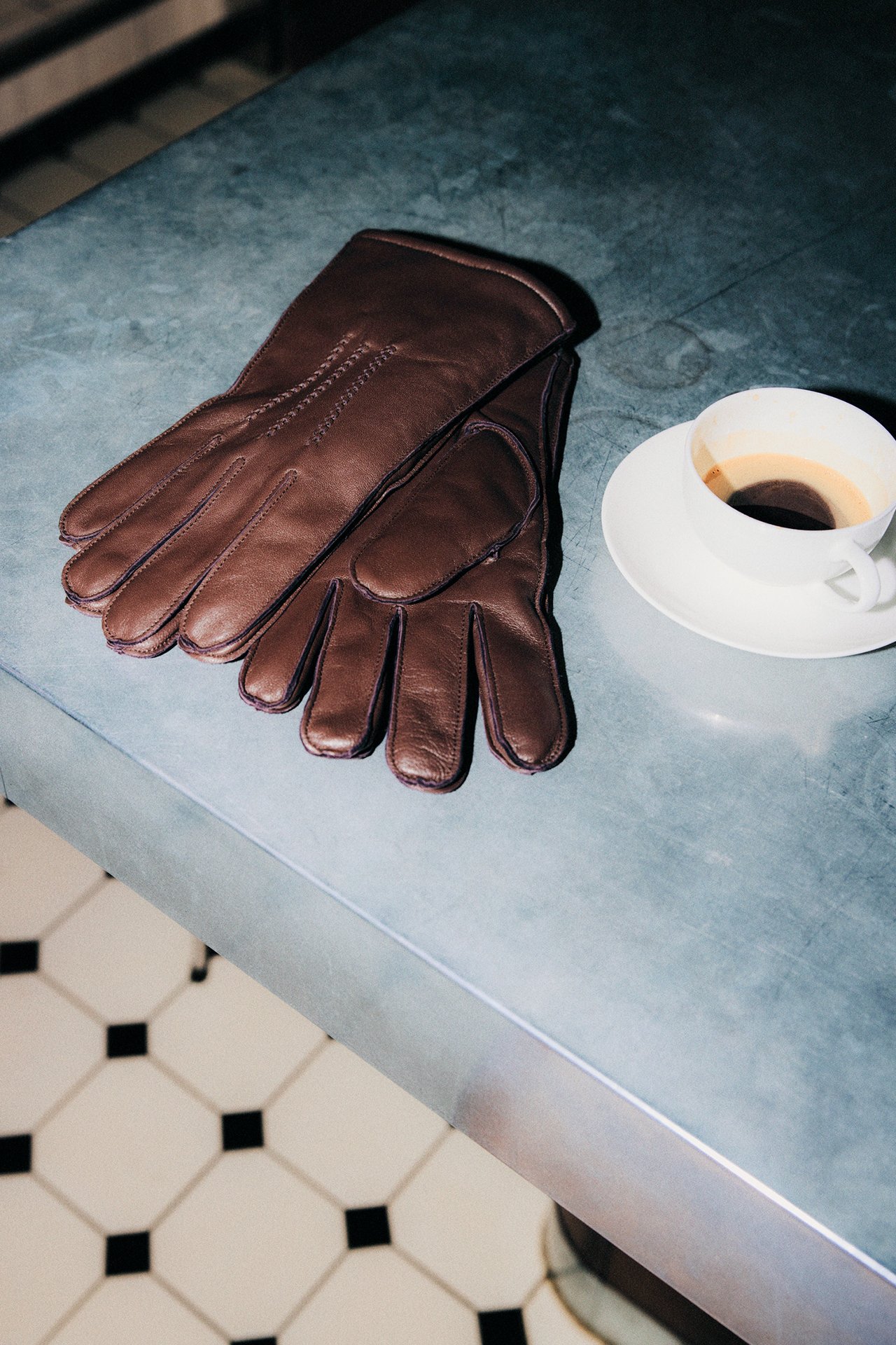 Nærbilde av brune skinnhansker med søm, ved siden av en hvit kopp kaffe på et fat.