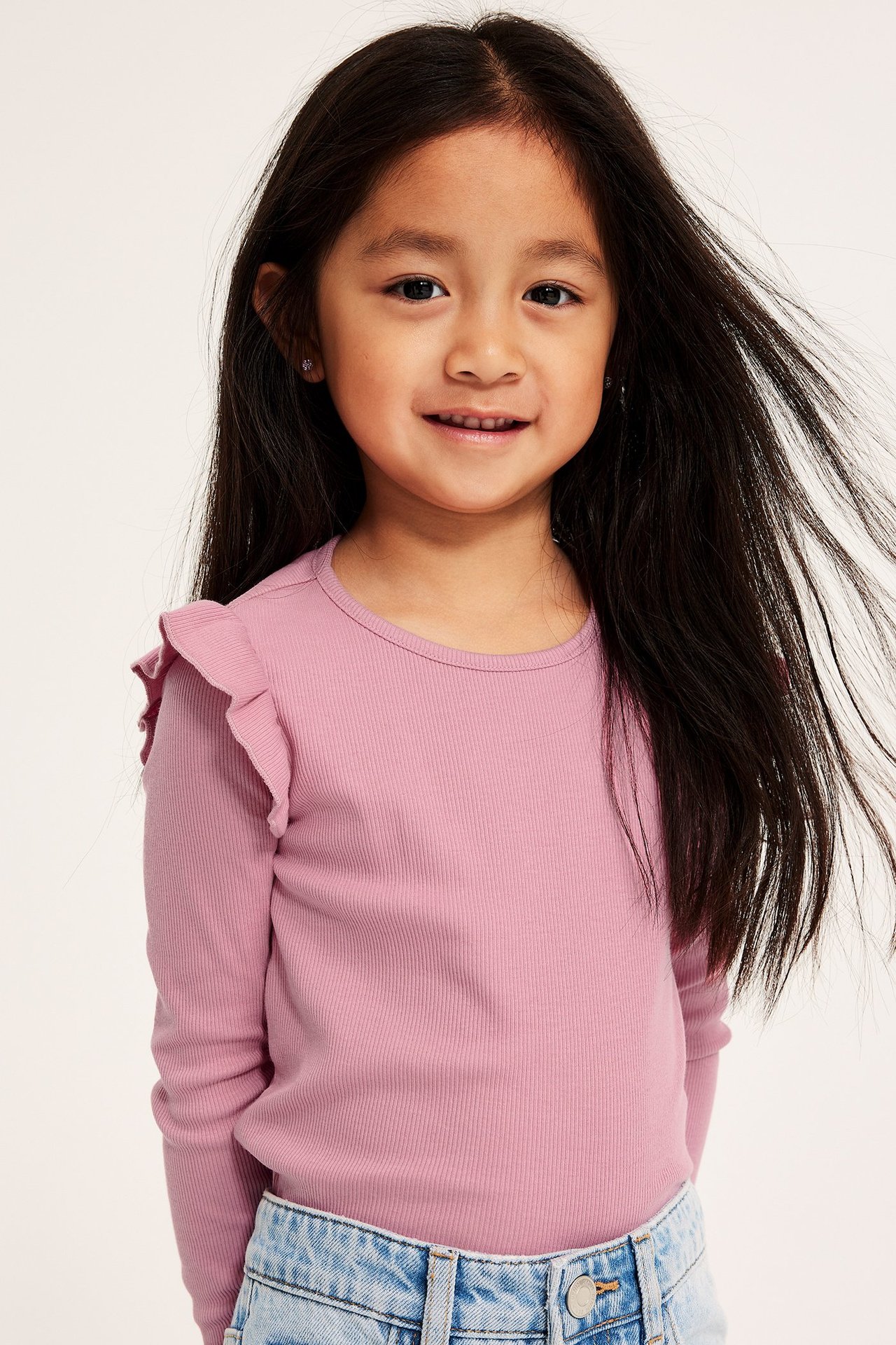 Frontvy av ett barn i rosa ribbad långärmad topp med volanger och ljusblå jeans.