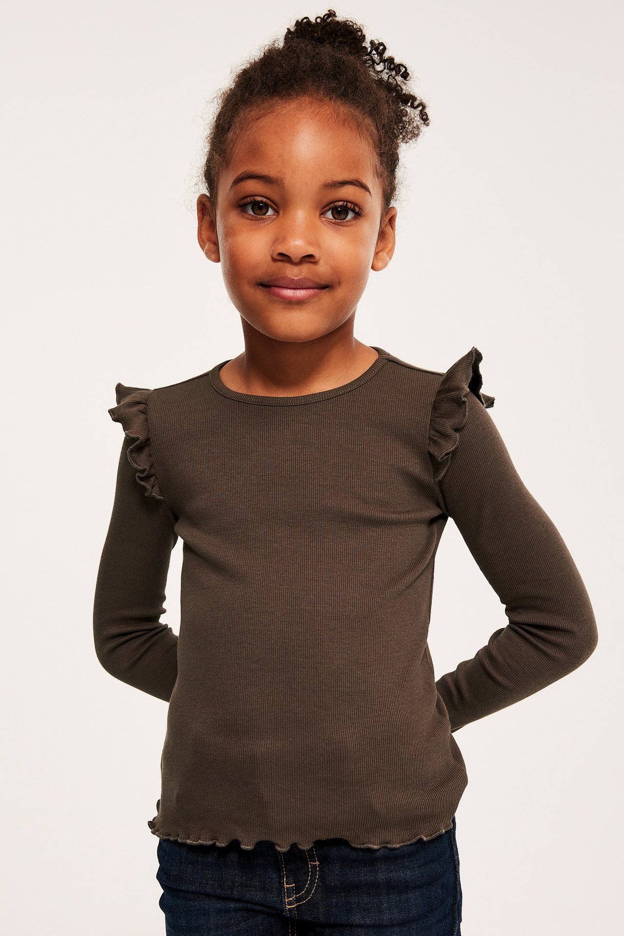 Edestä kuvattu lapsi yllään tummanruskea ribattu pitkähihainen toppi röyhelöillä ja tummansiniset farkut.