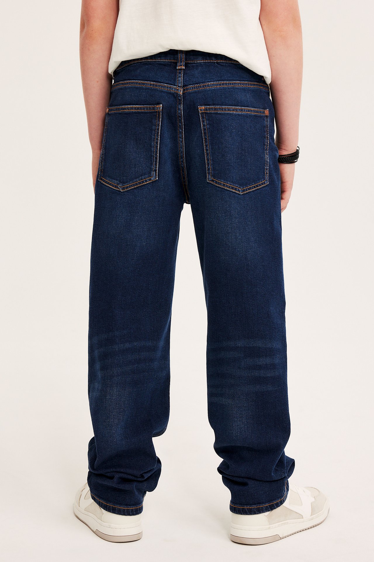 Bakfra av et barn i mørkeblå løse denimjeans, hvit t-skjorte, hvite joggesko og et svart armbånd.