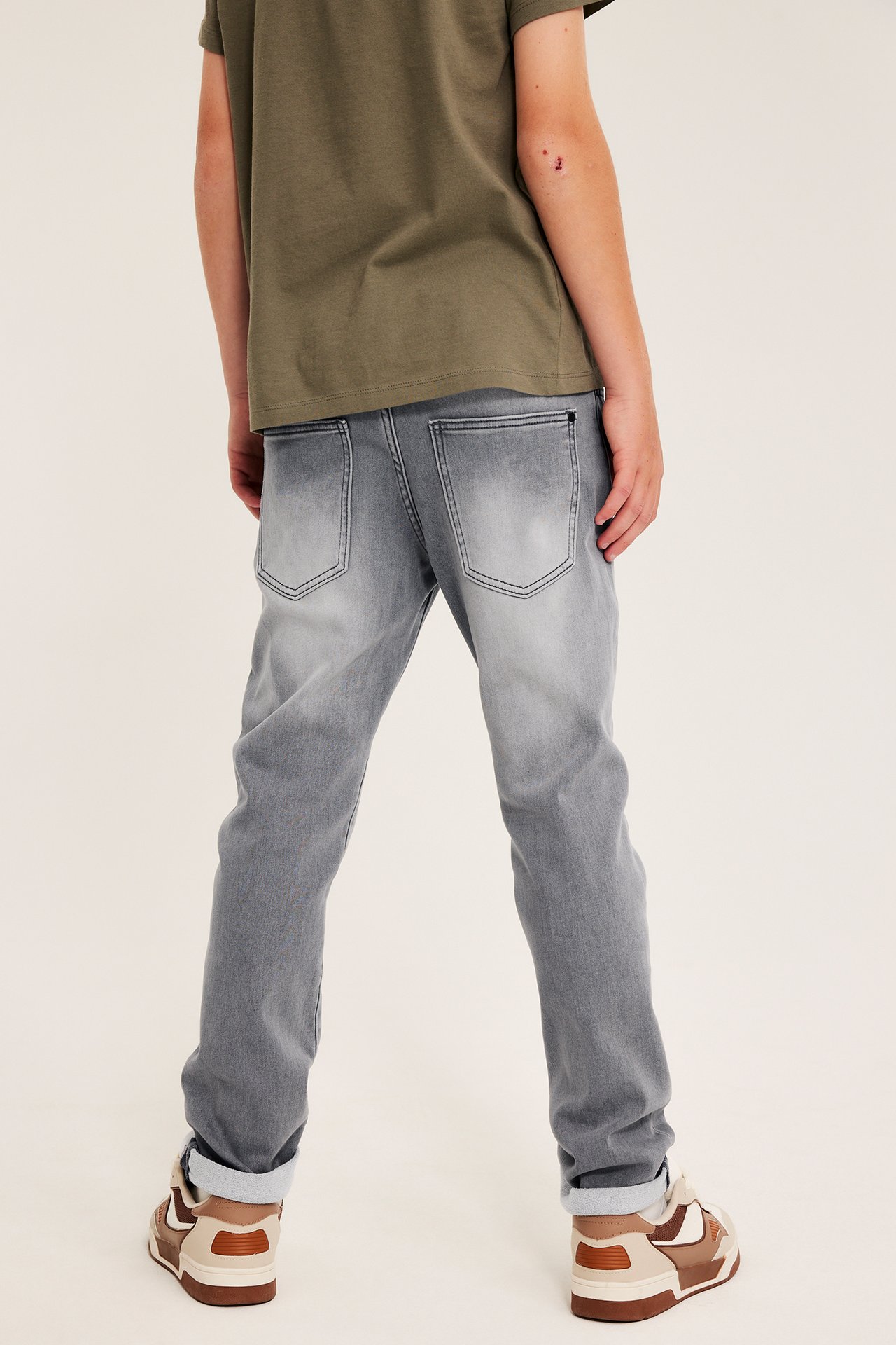Bakfra: Et barn i rette grå jeans, en olivengrønn t-skjorte og brune sneakers.