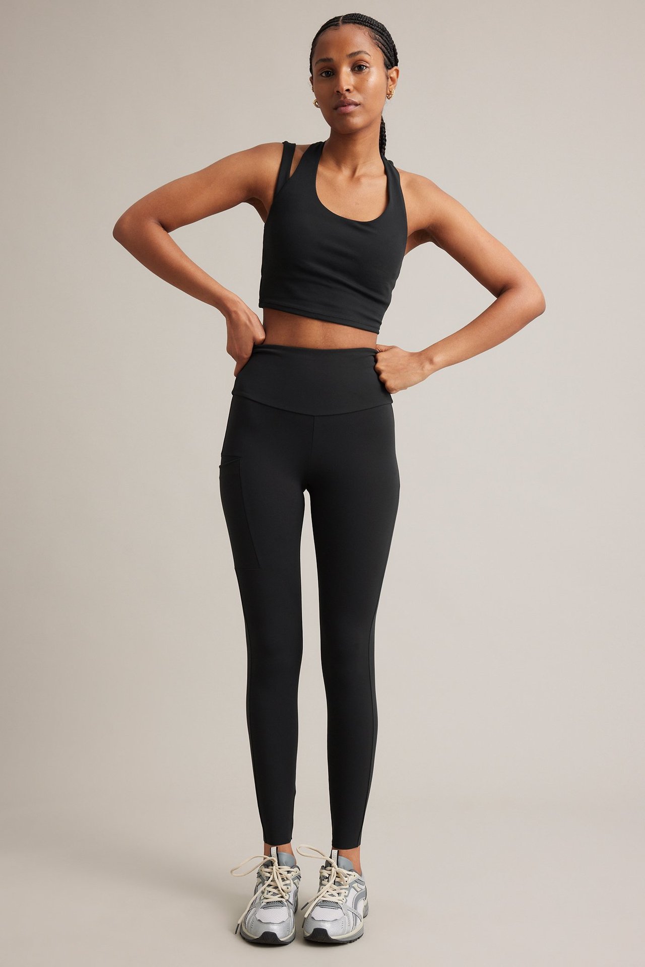Frontbilde av en kvinne i svart sportstopp med detaljerte stropper, svarte leggings og grå joggesko.