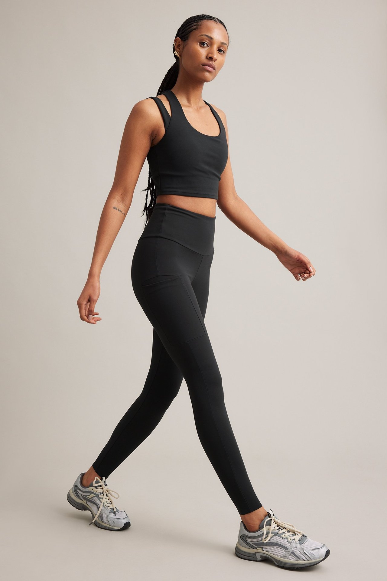 Sidebilde av en kvinne i svart kort sportstopp, svarte leggings med sidelommer og grå joggesko.