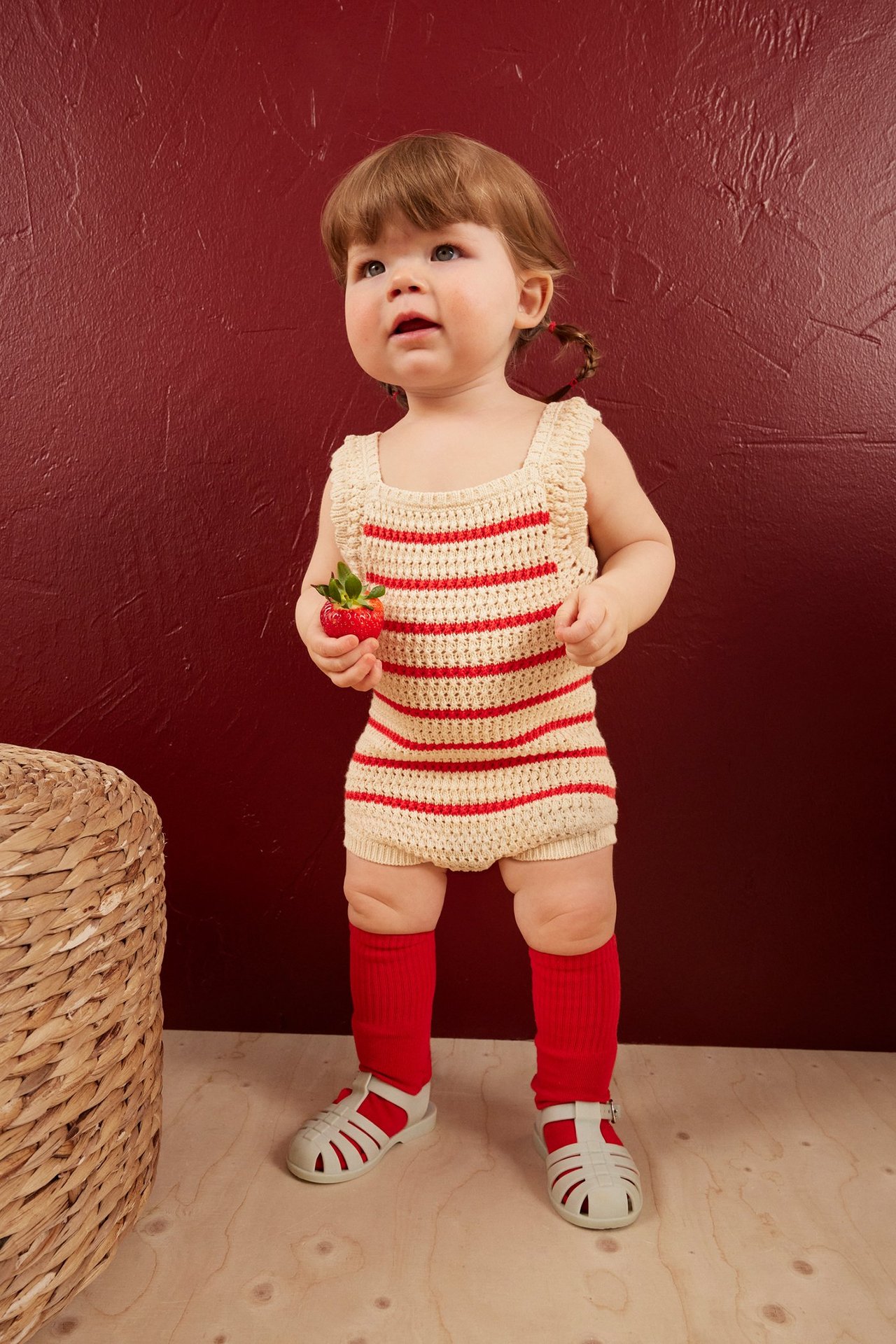 Etunäkymä: Vauva beigessä, punaraidallisessa neulotussa bodyassa, punaisissa sukissa ja harmaissa sandaaleissa, pitäen mansikkaa.