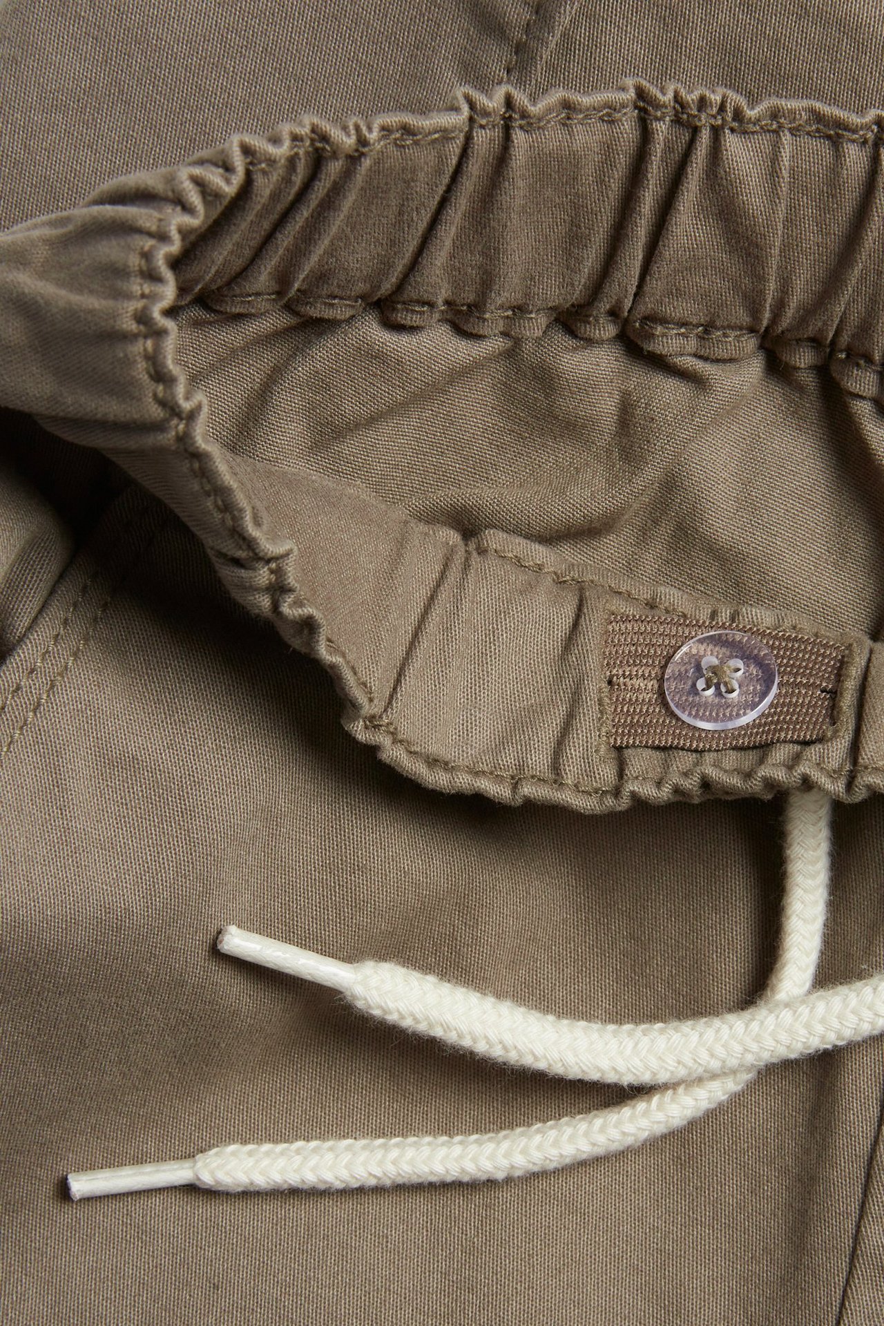 Nærbilde av brune cargobukser i bomull med elastisk linning, hvit snøring og innvendig justerbar knapp.