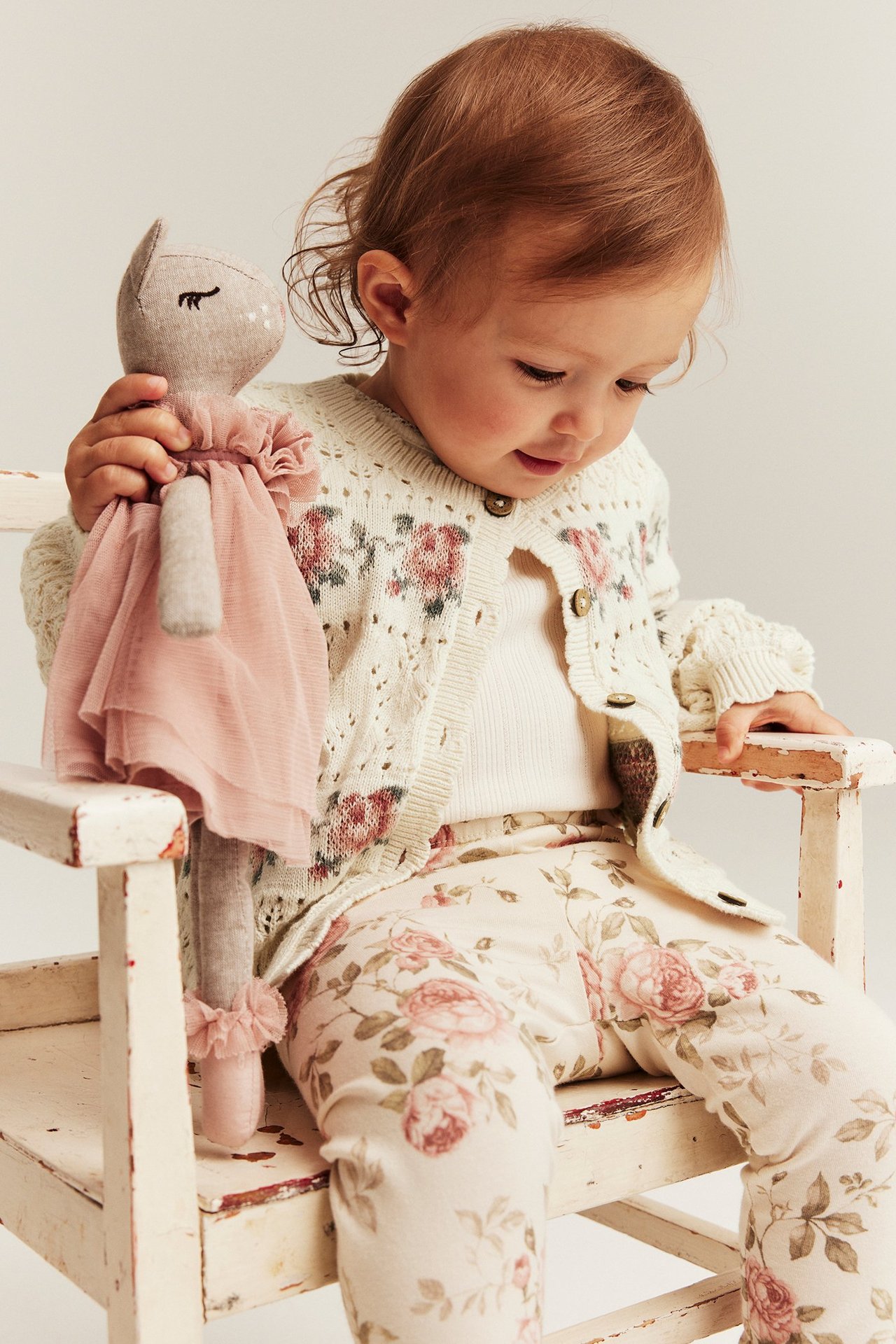 Vauva luonnonvalkoisessa jacquard-neuletakissa ruusuilla, valkoinen toppi, kukkaiset leggingsit, pitää nukkea.