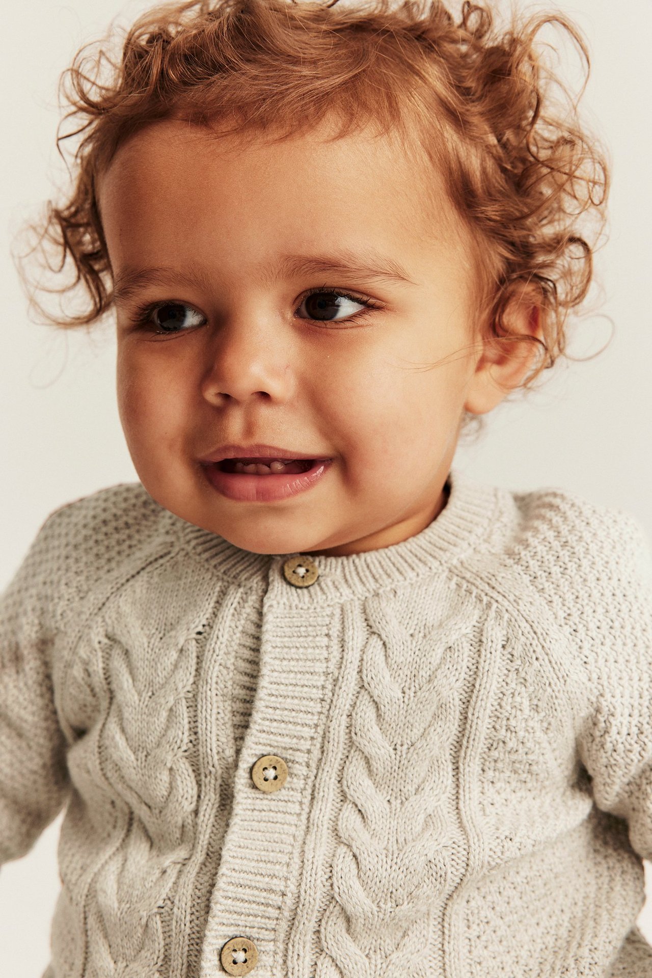 Lähikuva edestä vauvasta, jolla on beigemelange-värinen palmikkoneuleinen neuletakki puunapeilla.
