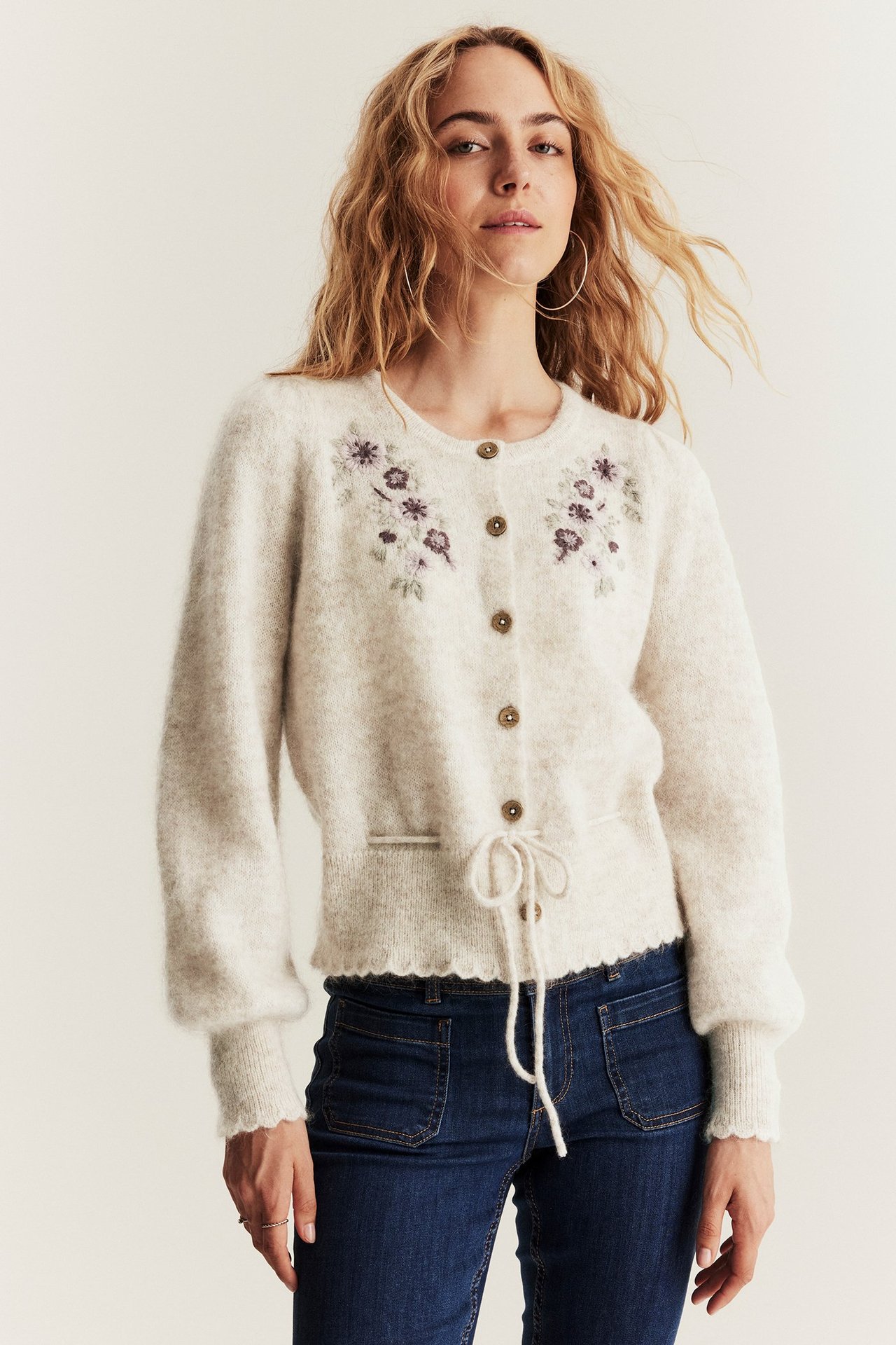 Framifrån: Kvinna i beige ullmixkofta med blommig brodyr, knappar, dragsko och mörkblå jeans.