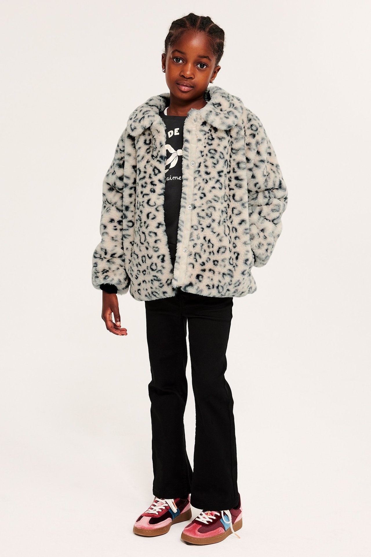 Framifrån: Barn i öppen beige leopardmönstrad fuskpälsjacka, svart t-shirt, svarta byxor, färgglada sneakers.