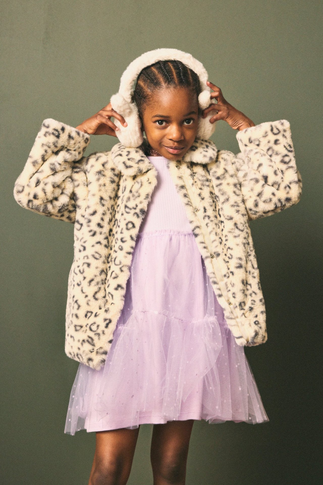 Widok z przodu dziecka w beżowej kurtce ze sztucznego futra w panterkę, liliowej sukience i beżowych nausznikach.