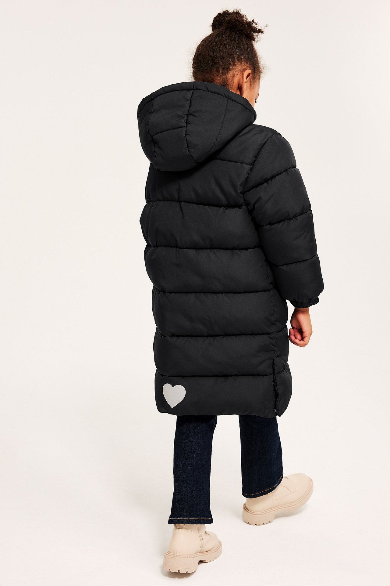 Bakfra: Lang svart pufferjakke for barn med hette, reflekterende hjerte, mørke jeans og lysebeige støvler.