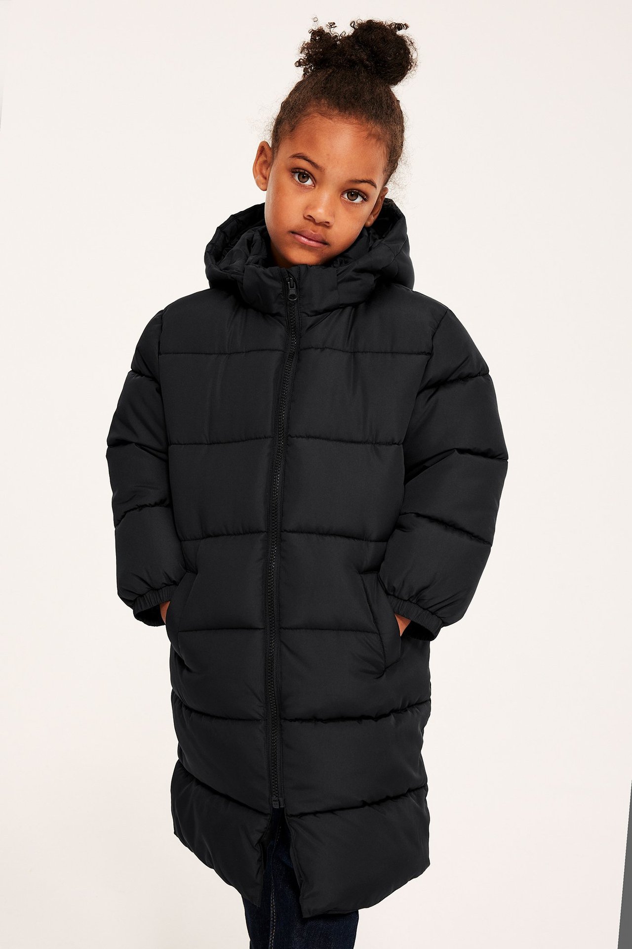 Framifrån: Barn bär lång svart vadderad jacka med huva, dragkedja, fickor, över mörka jeans.