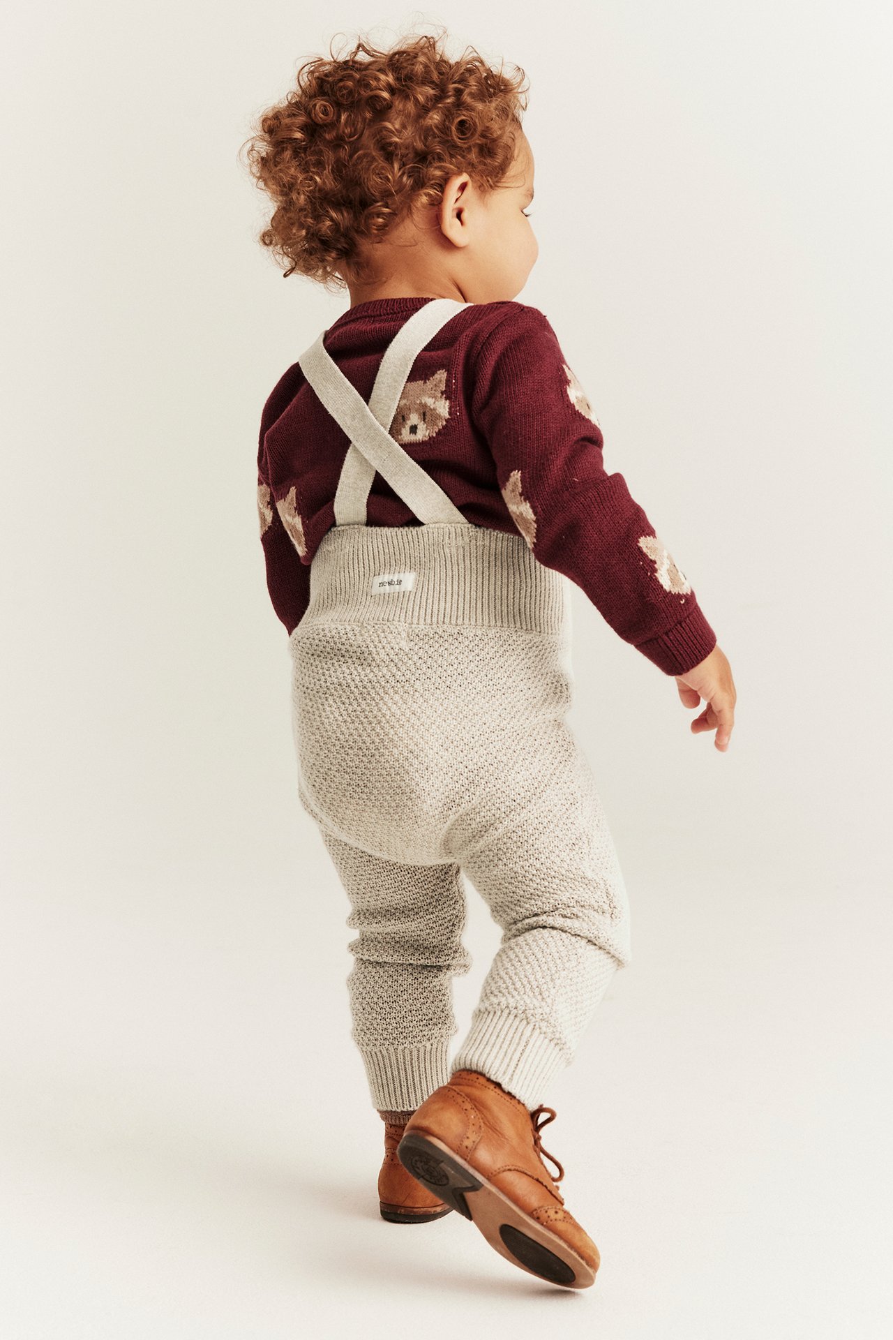 Bakifrån: baby i beige melange stickade hängselbyxor över en vinröd tröja med björnmönster och bruna skor.