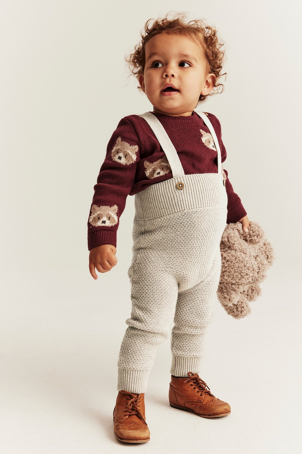 Frontbilde av en baby i beige strikket selebukse, burgunderfarget dyregenser, brune sko, holder en bamse.