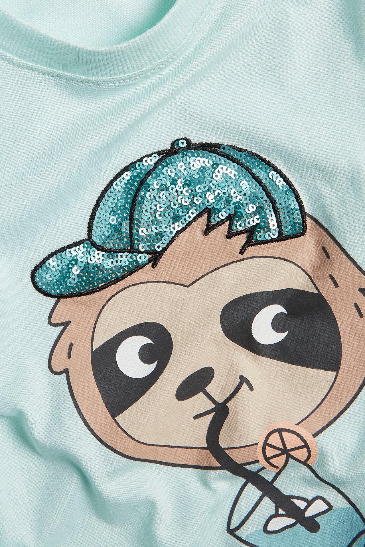 Zbliżenie na jasnoturkusowy bawełniany T-shirt dziecięcy z nadrukiem leniwca w cekinowej czapce.