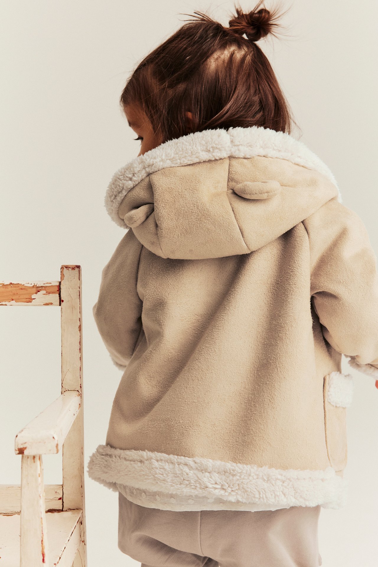 Widok z tyłu: Dziecko w beżowej kurtce barankowej, białym futerkiem, kapturem z uszami i jasnych spodniach.