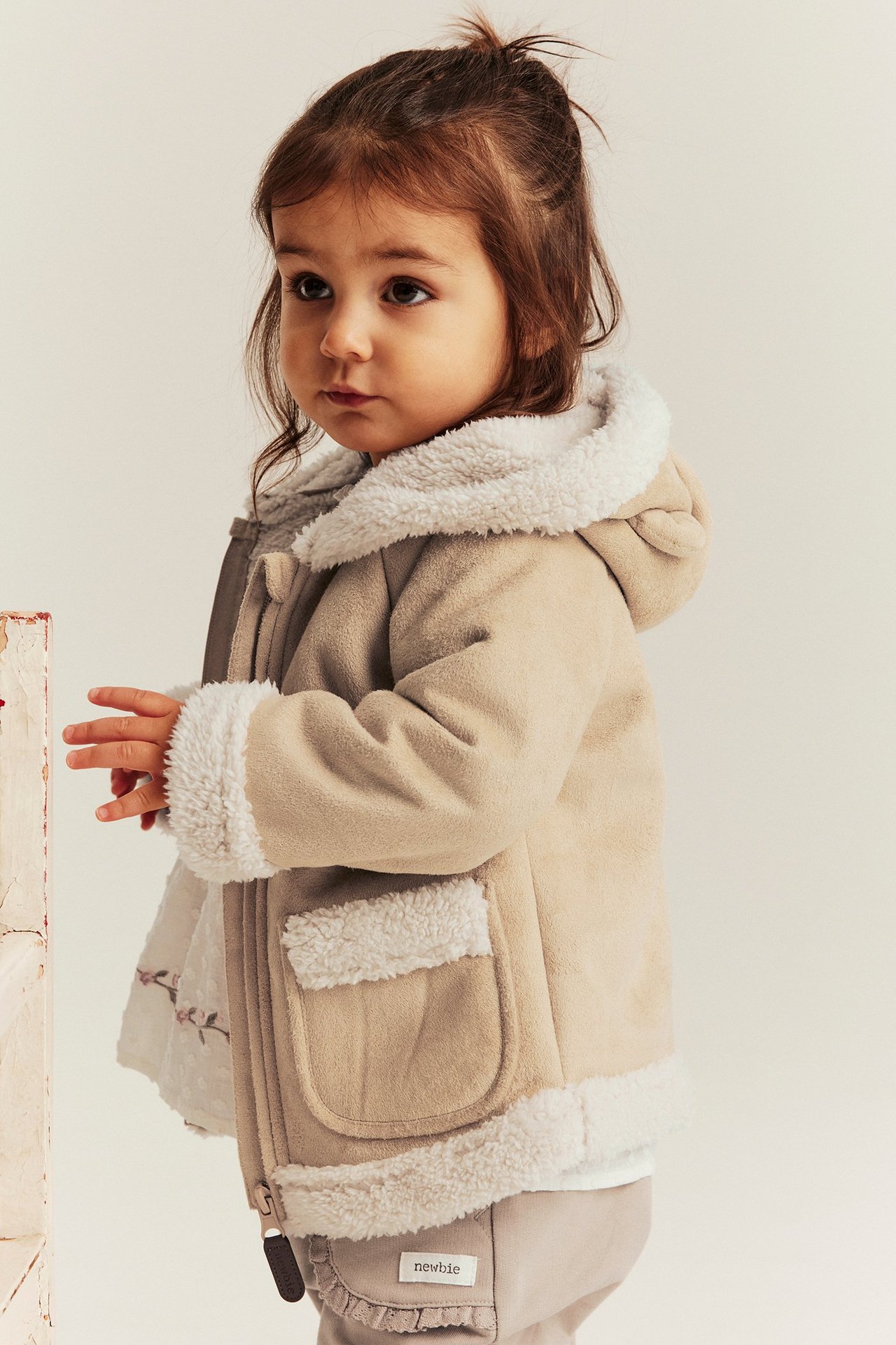 Front-sidevisning av et barn i beige shearling-jakke med kremfarget fôr, hette med bjørneører.