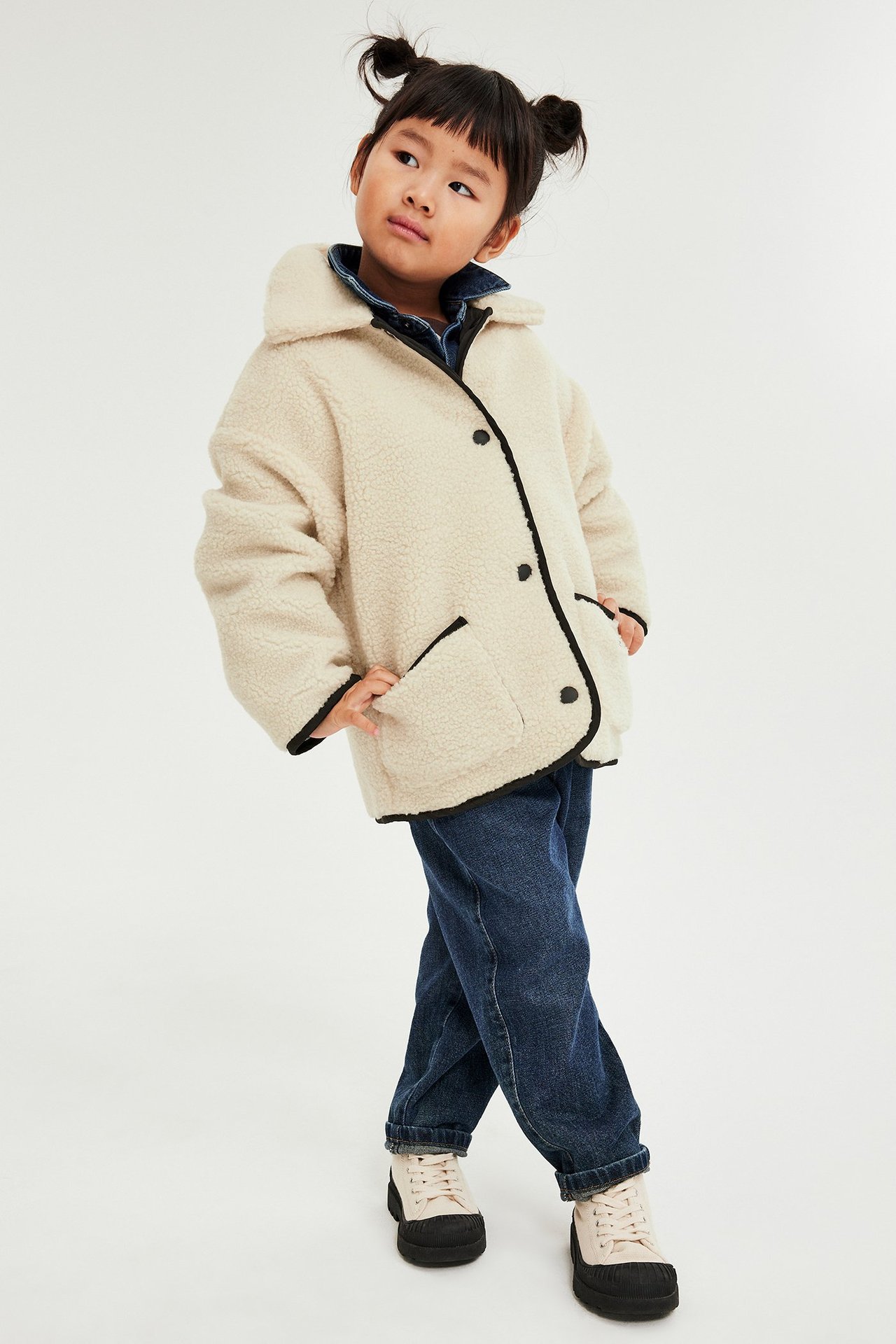 Barn i beige pilejakke med sorte kanter, blå dongeriskjorte, mørkeblå jeans og beige sko.