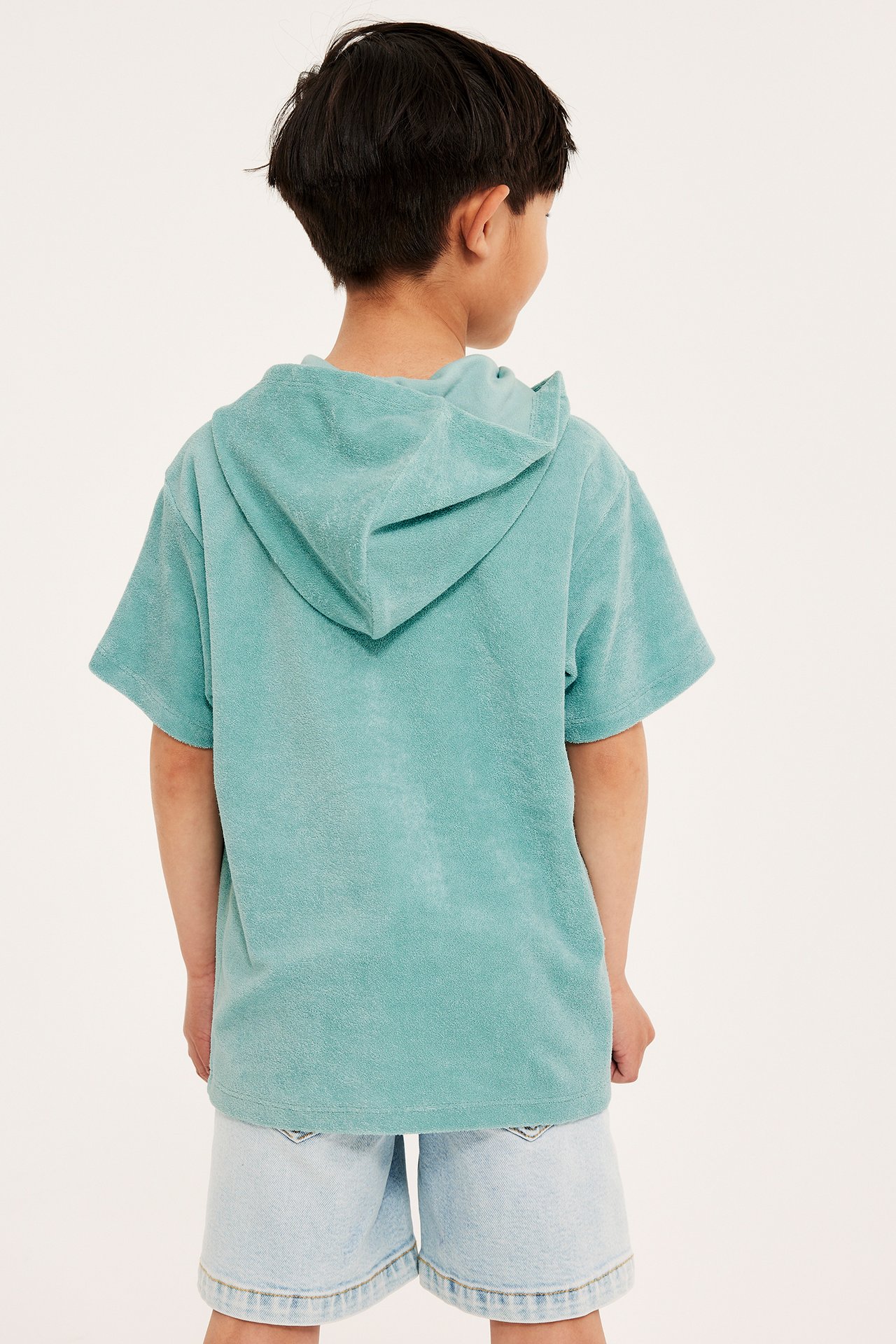 Bakifrån: ett barn i en oversized ljusgrön kortärmad frottéhoodie och ljusblå denimshorts.