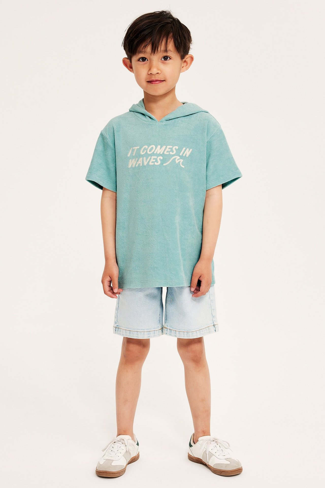 Framifrån: Barn i ljusgrön oversized frottéhoodie med brodyr, ljusblå shorts, vita sneakers.